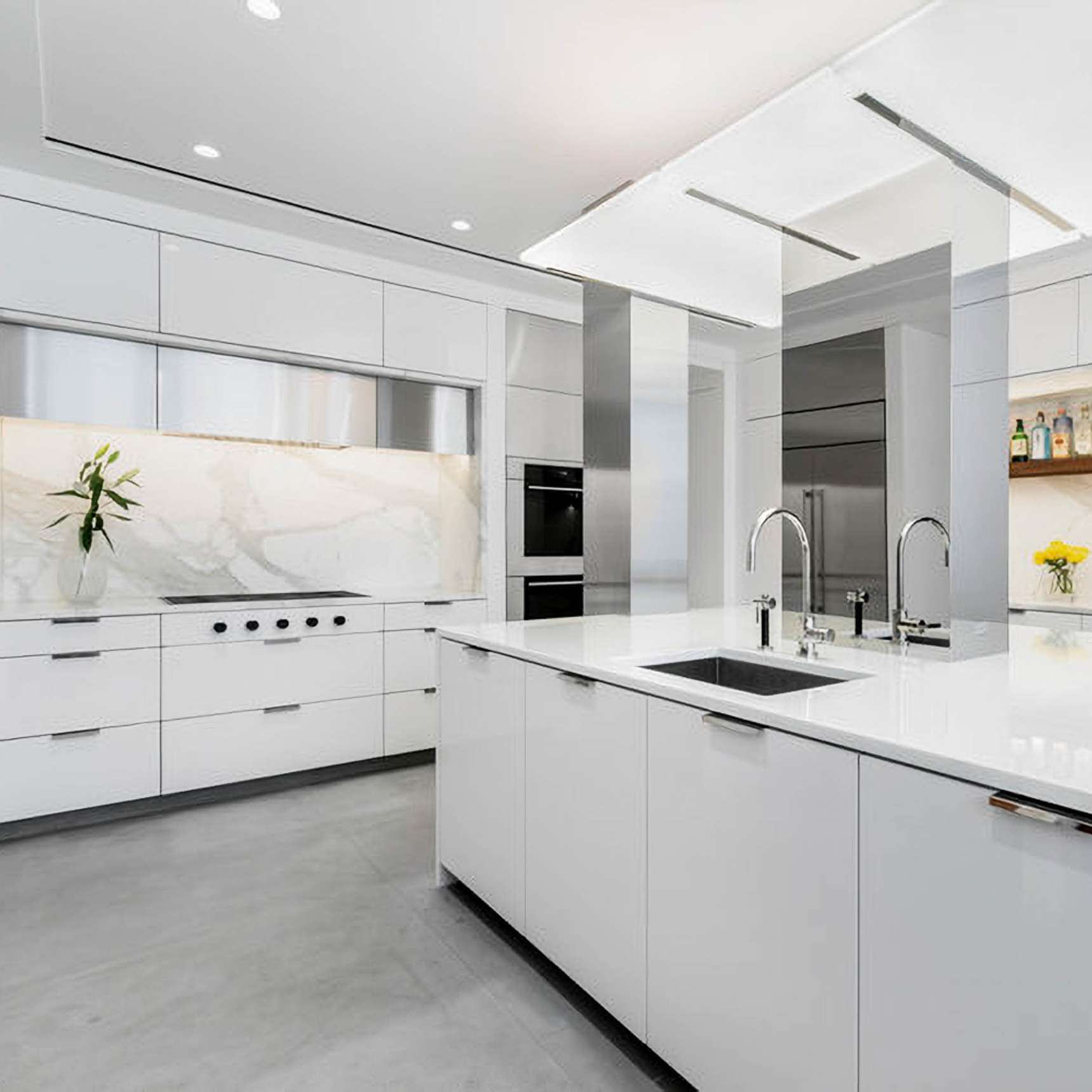 Modern white kitchen with marble backsplash, center island with built-in sink, stainless steel appliances, and recessed lighting.