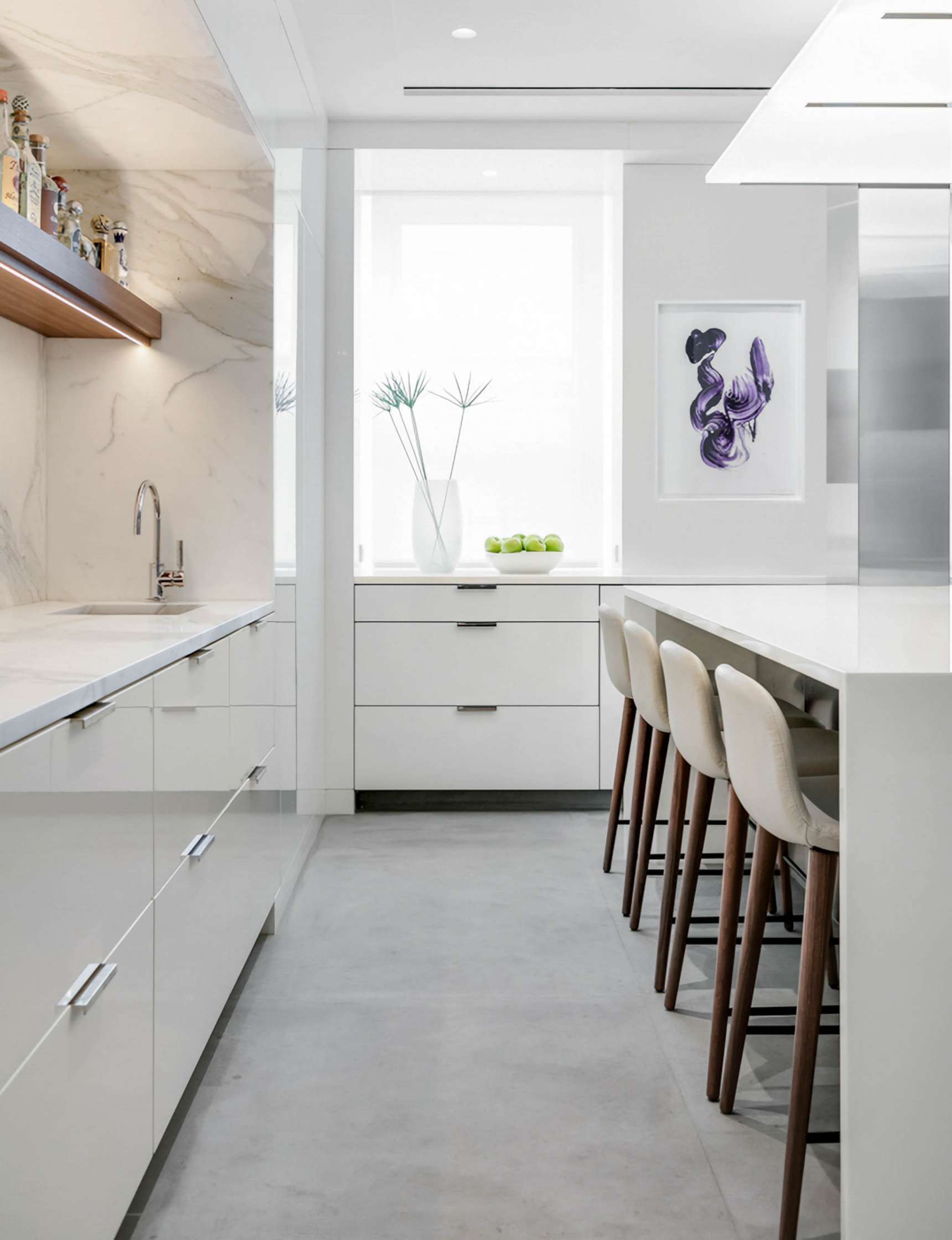 A modern kitchen with white cabinets, a marble backsplash, a row of bar stools along an island, and a vase with tall stems on the windowsill. A framed art piece and a bowl of green apples are also visible.