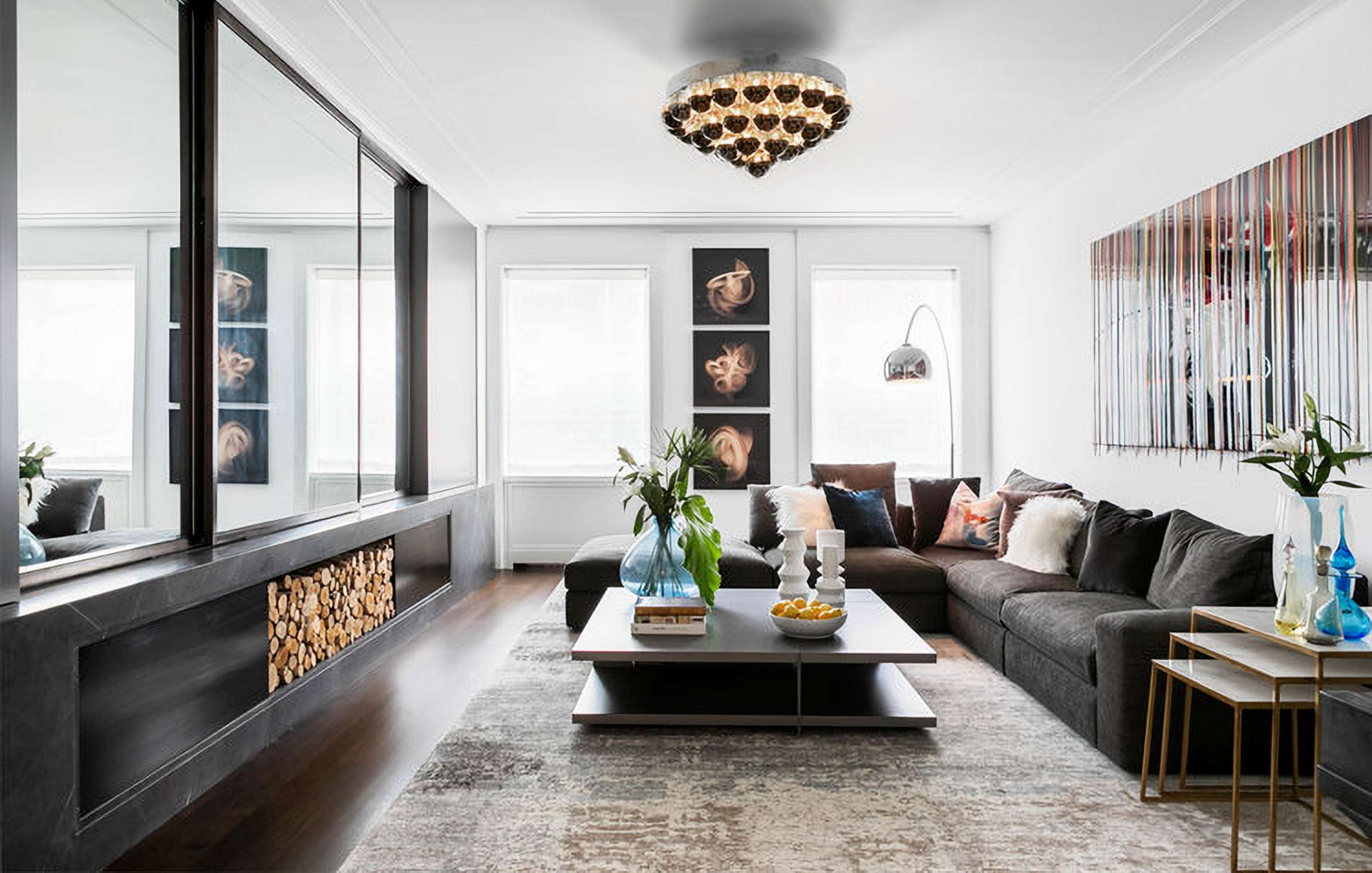 Modern living room with a sectional sofa, coffee table, contemporary artwork, and a wall-mounted fireplace. A chandelier hangs from the ceiling, and there are large windows allowing natural light.