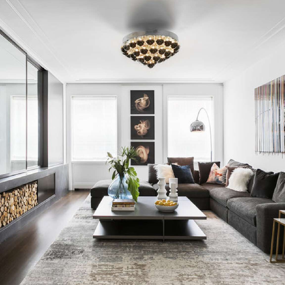 Modern living room with a large sectional sofa, a central coffee table, and decorative items. The room features framed art on the wall, a unique ceiling light fixture, and a built-in wood storage area.