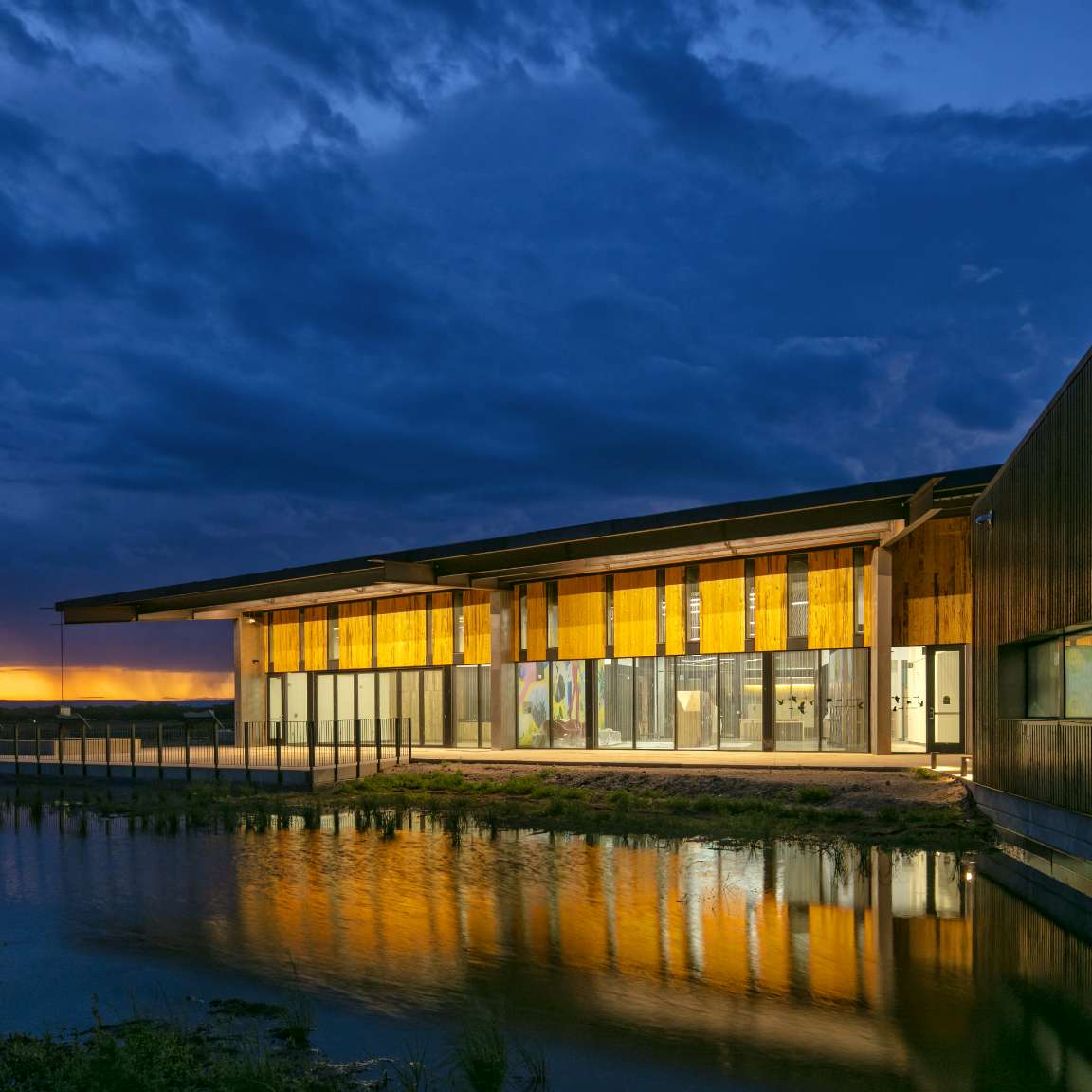 A modern building with large glass windows is illuminated against a twilight sky, reflecting on the still water in front of it.