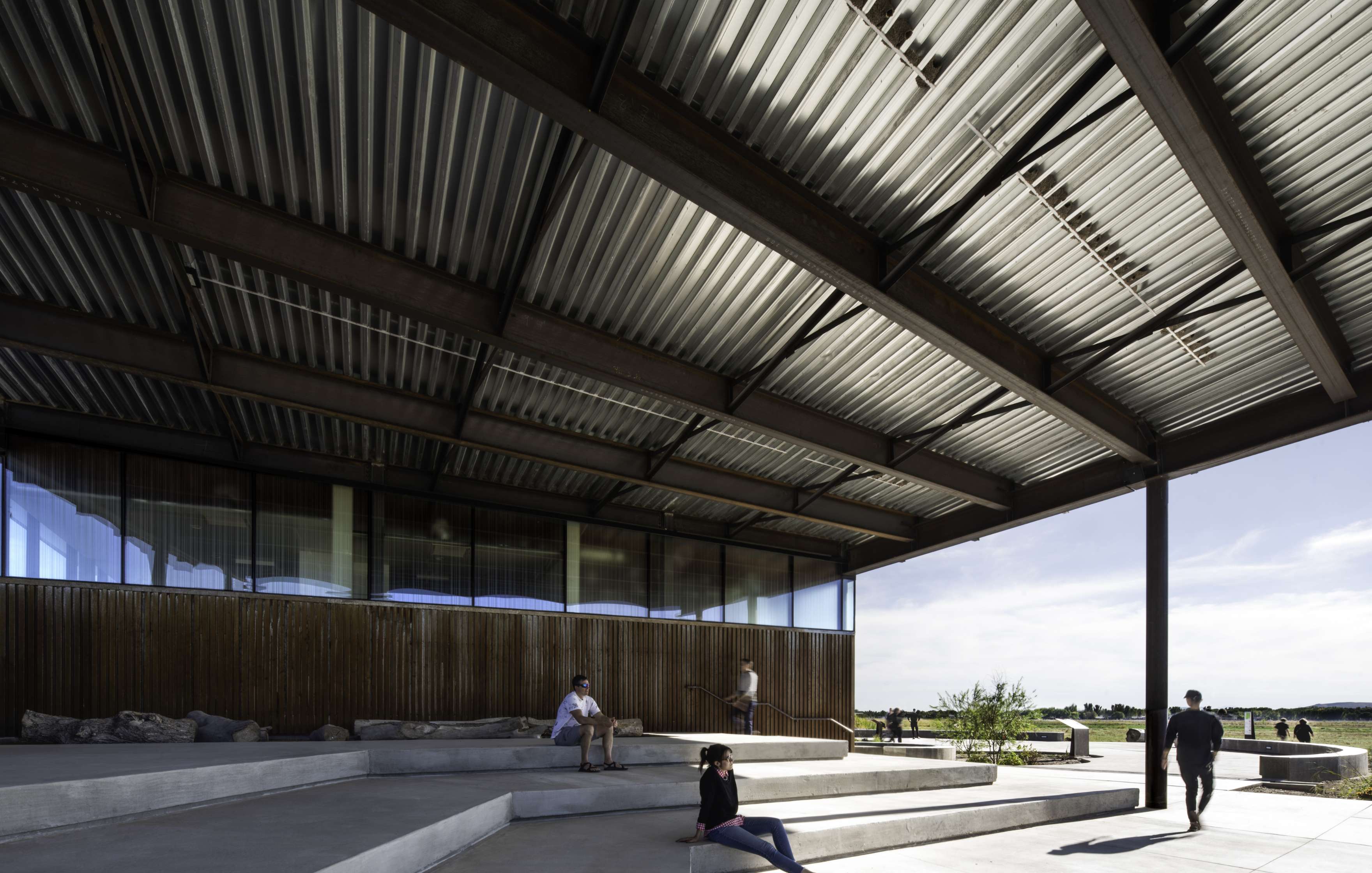 Several people sit and stand on tiered concrete steps beneath a large, corrugated metal canopy at an outdoor public space.