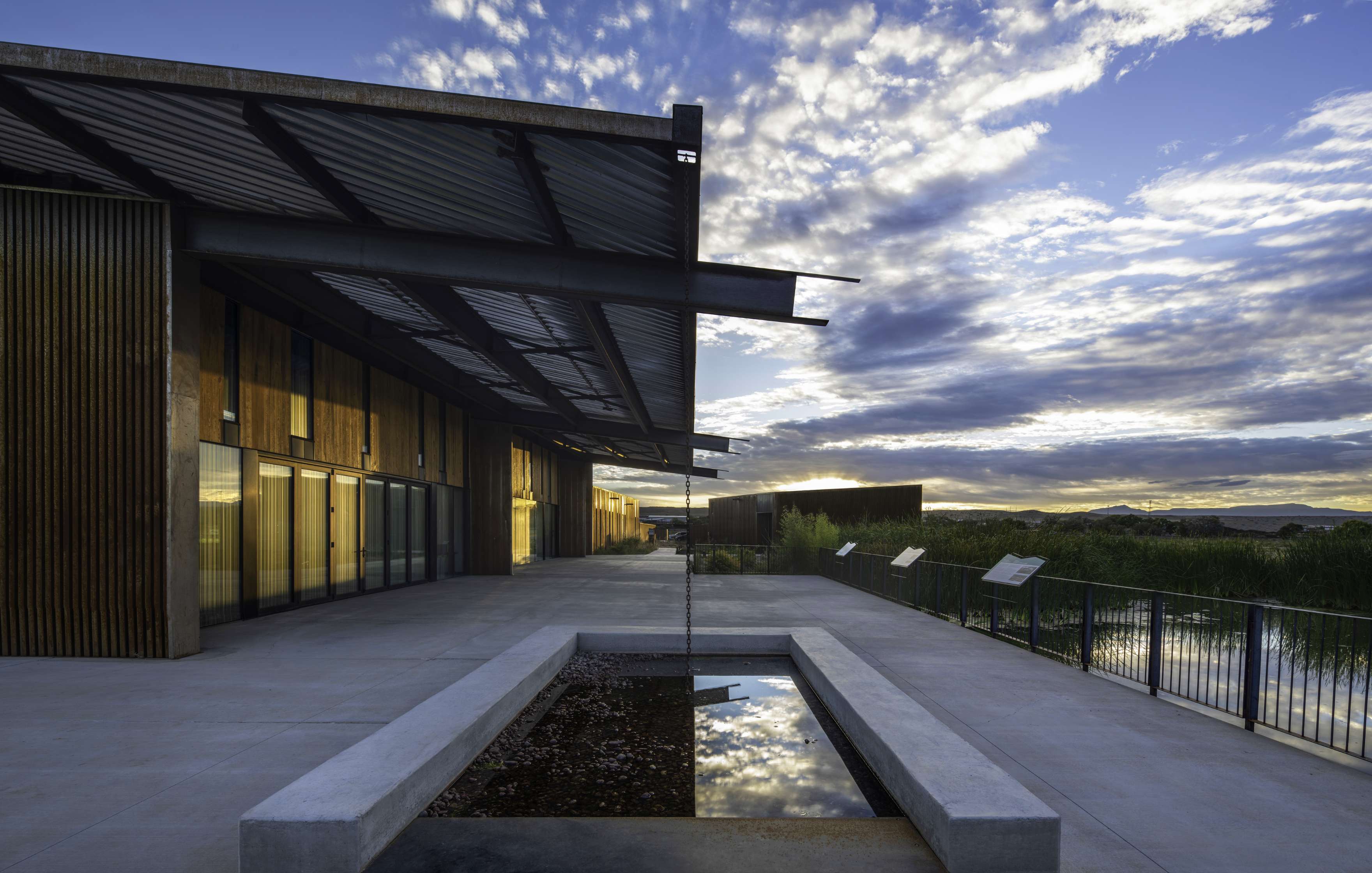 Contemporary building with large glass windows and a modern metal roof. A rectangular water feature is in the foreground, reflecting the cloudy sky during sunset.