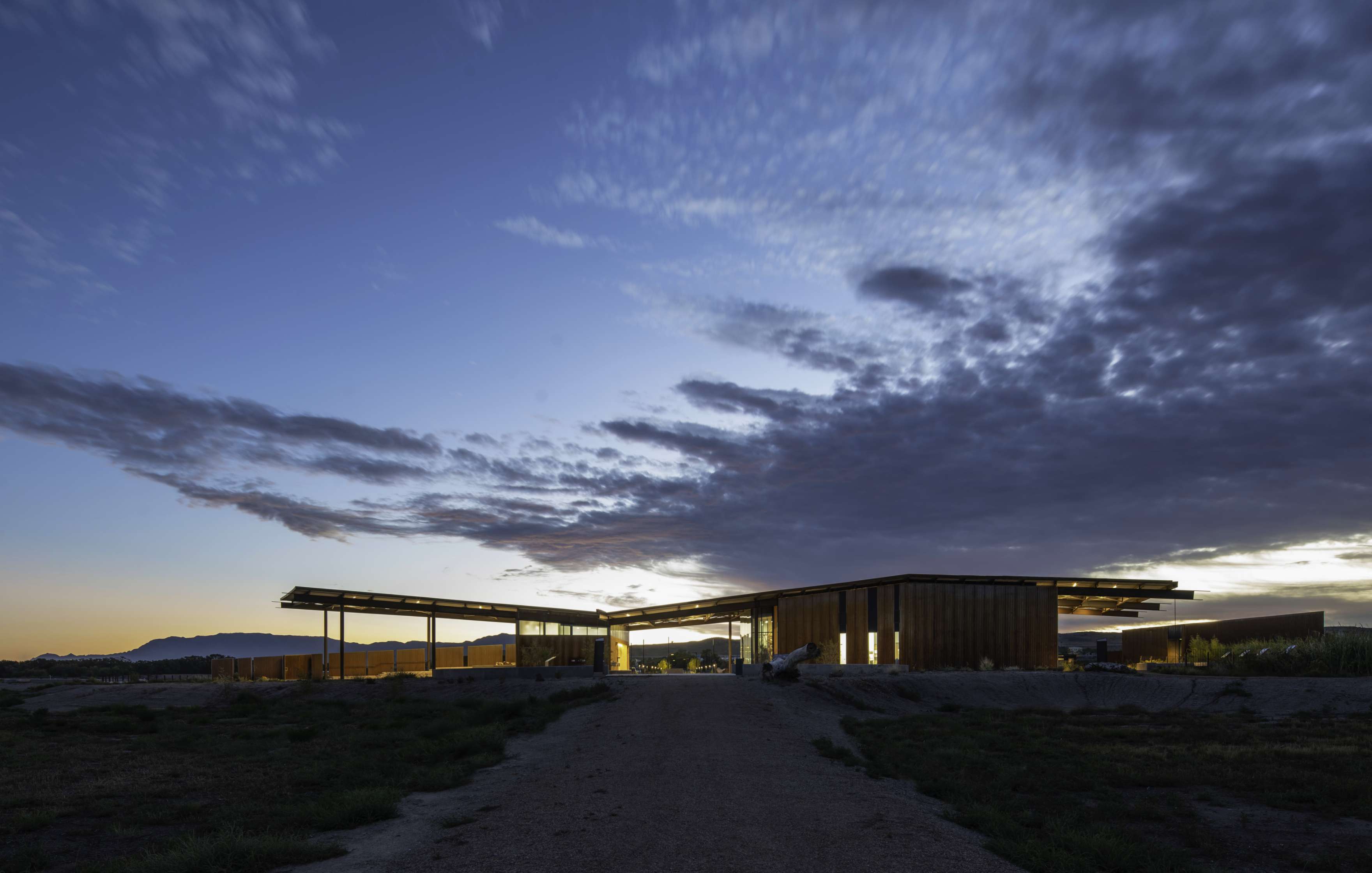 A modern building set against a dramatic dusk sky with scattered clouds. The structure features clean lines and substantial use of glass and steel, illuminated by interior lights.