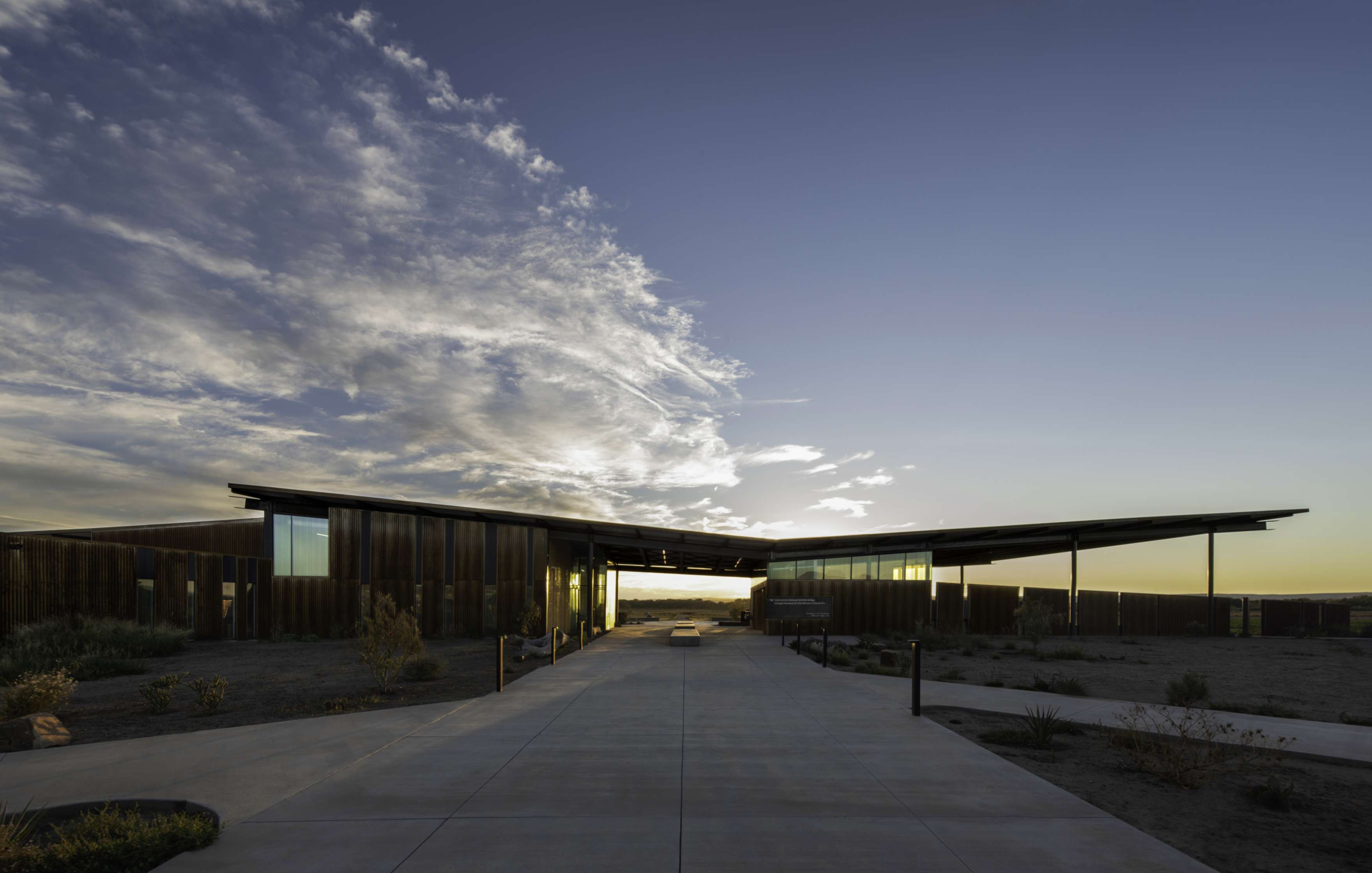 Modern building with a flat roof and large windows, set in a desert landscape under a wide sky, with a concrete pathway leading to the entrance.