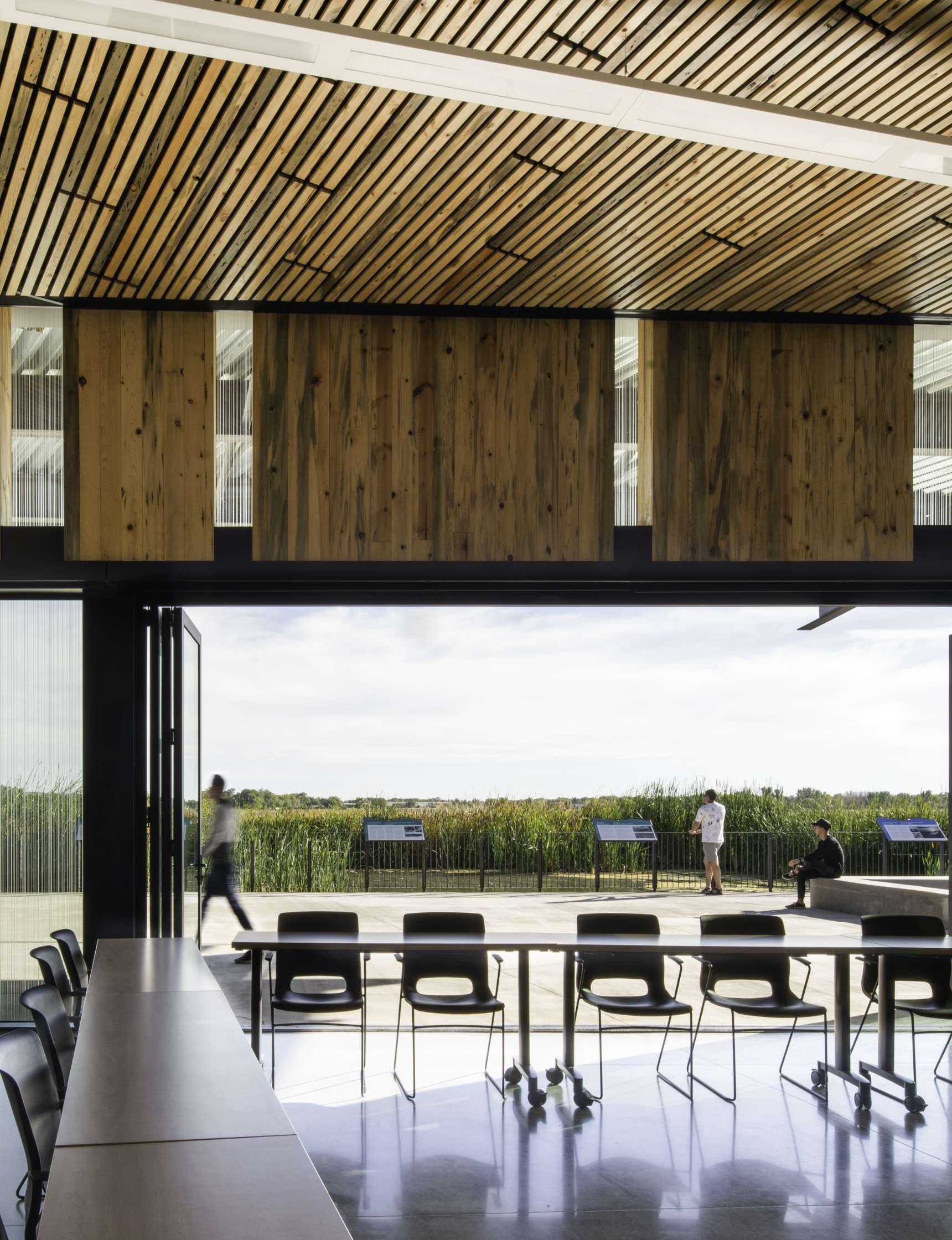 A modern indoor space with wooden ceiling panels and large windows overlooks an outdoor area with people standing and sitting. Black chairs and tables are arranged in the foreground.