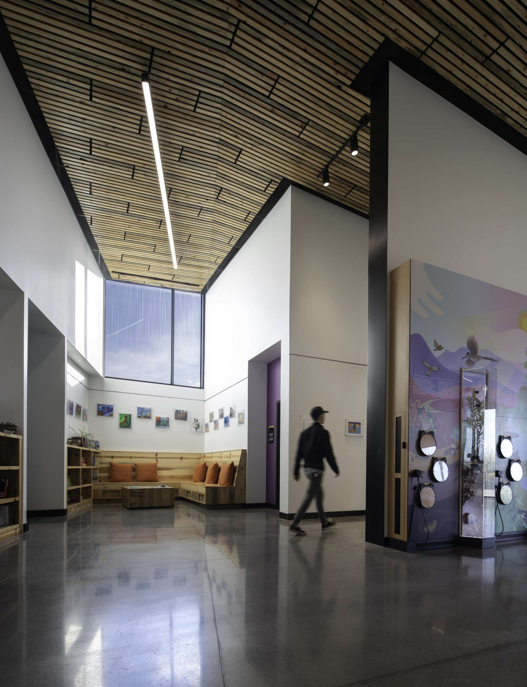 A spacious modern lobby with an intricate wooden ceiling and a mix of natural and artificial lighting. There are art pieces on the walls, seating with cushions in the corner, and a person walking by.