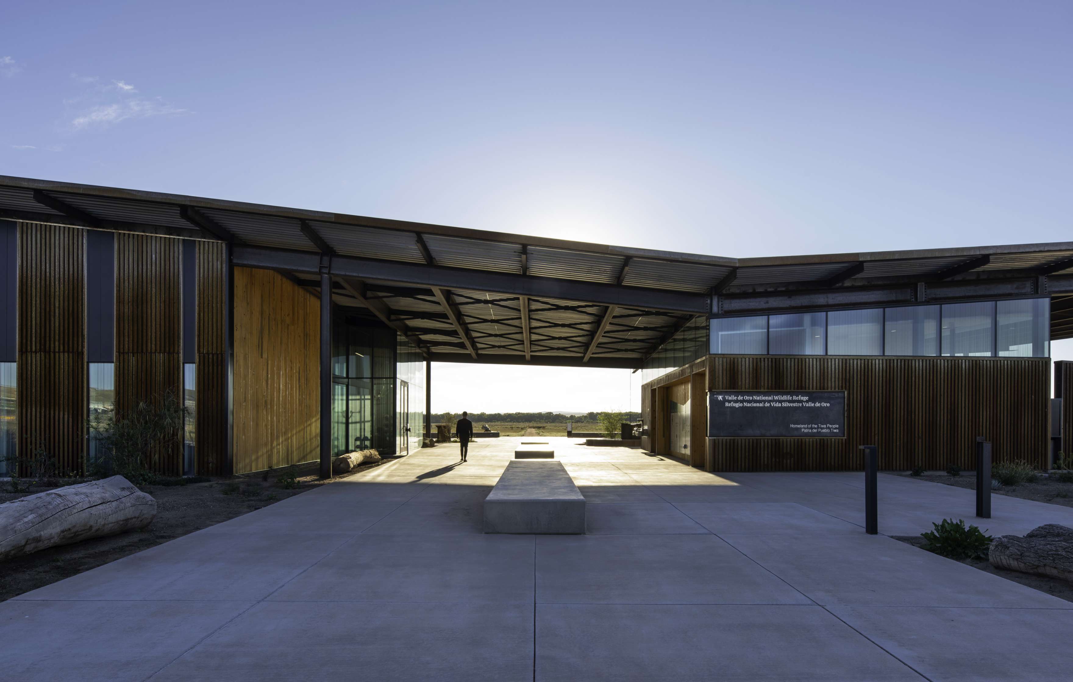 Modern building with large overhangs, glass walls, and wooden accents, situated under a clear sky. Sunlight shines from behind, casting shadows on the concrete pathway leading to the entrance.