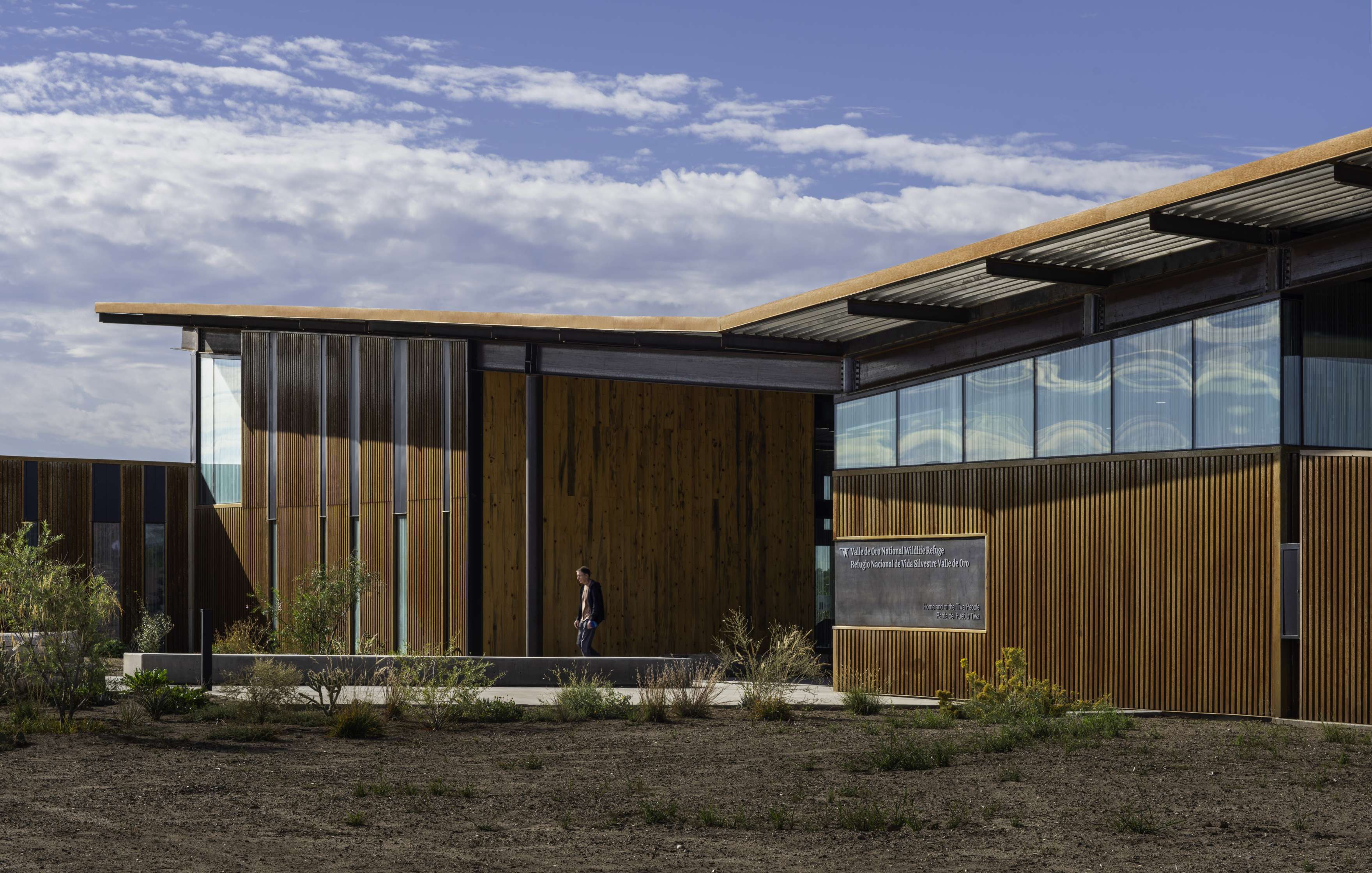 Modern building with wooden exterior panels and large glass windows, set against a partly cloudy sky. A person walks past the entrance. Sparse vegetation is visible in front of the building.