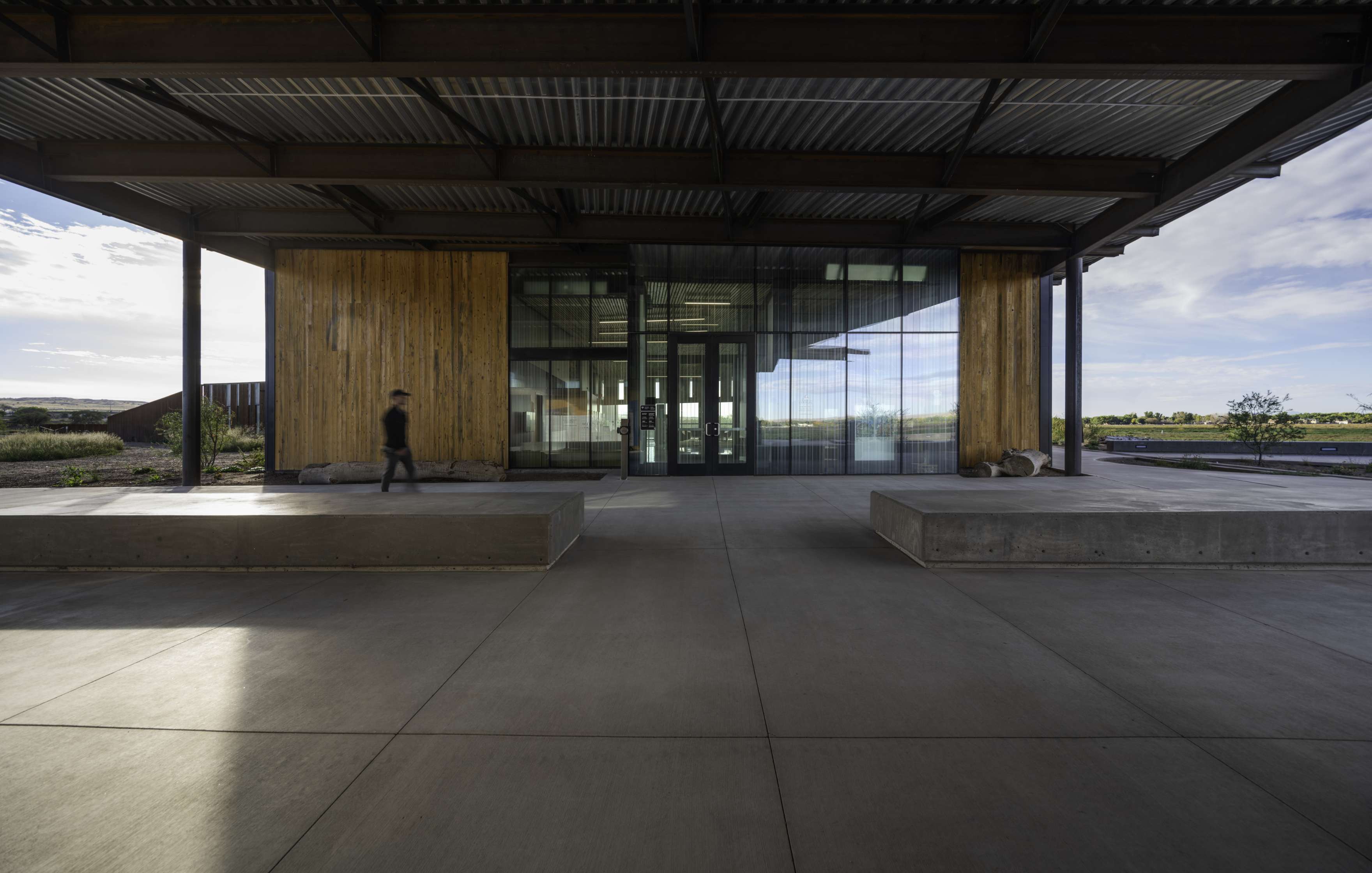 A modern building with large glass doors and wooden walls, featuring metal roofing. A person walks by, and concrete benches are positioned in front of the entrance.