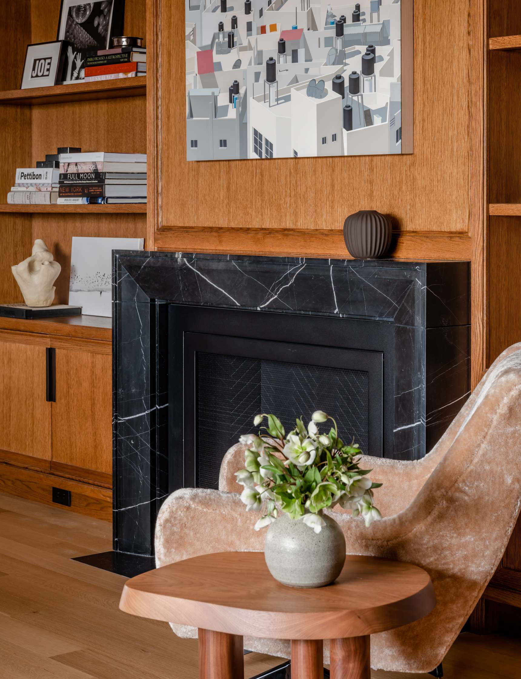 A cozy living room with a black marble fireplace, a vase with flowers on a wooden table, a plush velvet chair, and books and decorative items on wooden shelves. A cityscape artwork is above the fireplace.