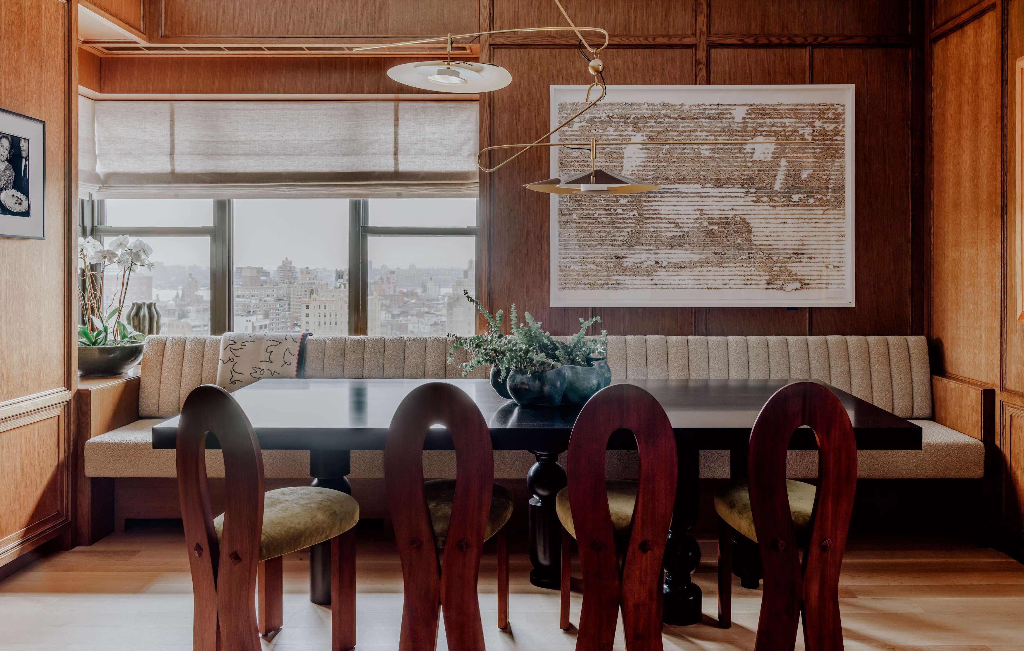 A dining area featuring a large rectangular table with five wooden chairs. The space includes a built-in cushioned bench, framed artwork, a window with roller shades, and a ceiling light fixture.