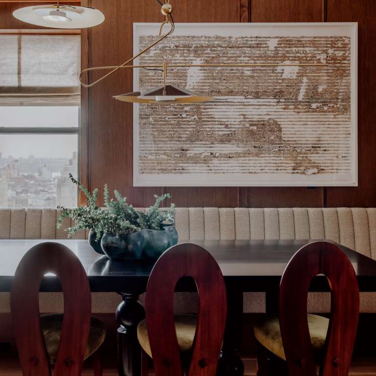 A dining area with wooden walls and floor, featuring a rectangular table and six chairs. The space is decorated with a large painting and potted plants, leading to a brightly lit living room.