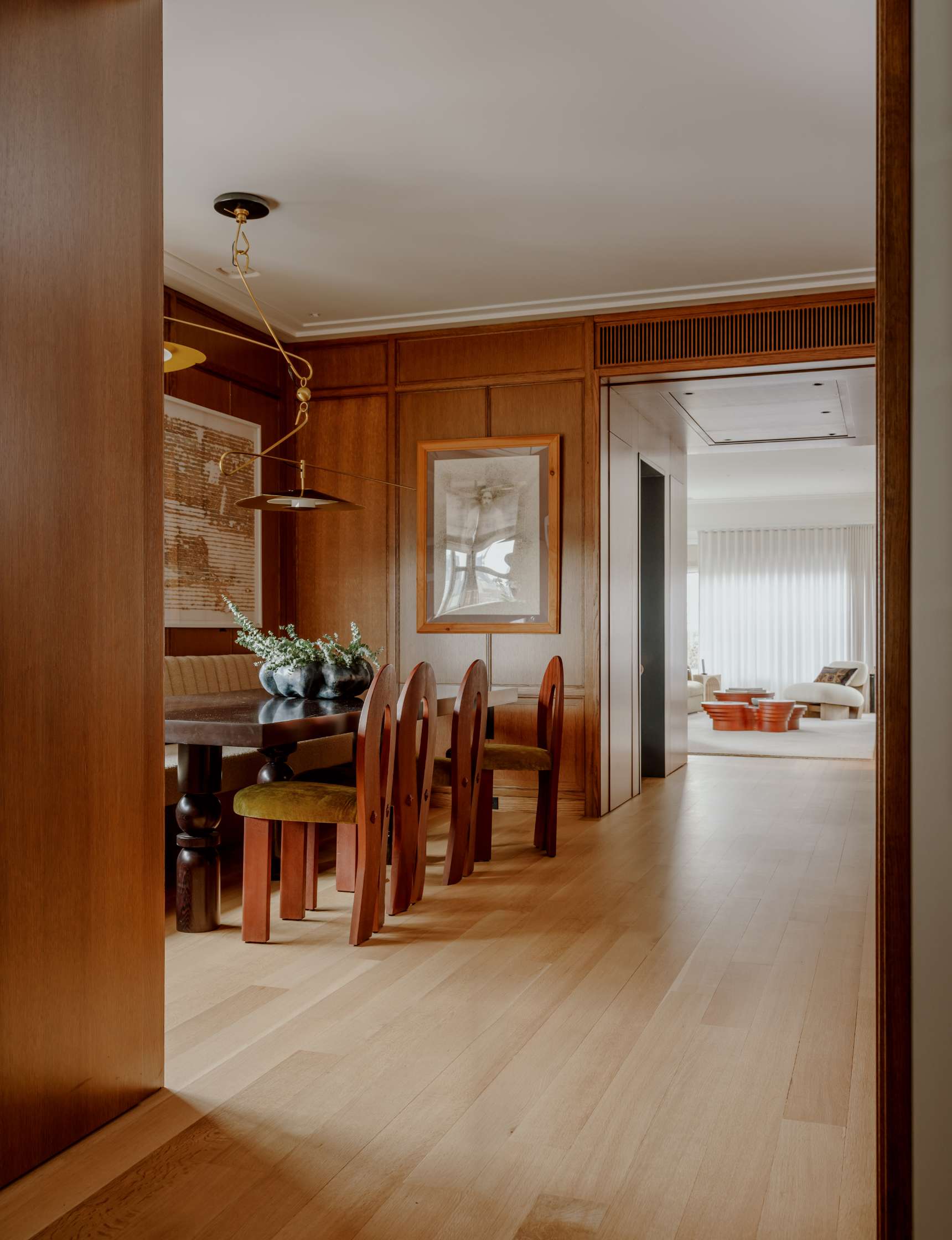 A dining area with wooden walls and floor, featuring a rectangular table and six chairs. The space is decorated with a large painting and potted plants, leading to a brightly lit living room.