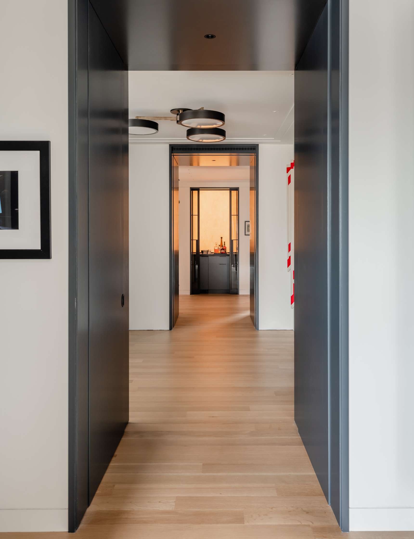 A hallway with light wooden floors leads to an open doorway with a room featuring a black cabinet and overhead lighting. A framed picture is on the left wall.