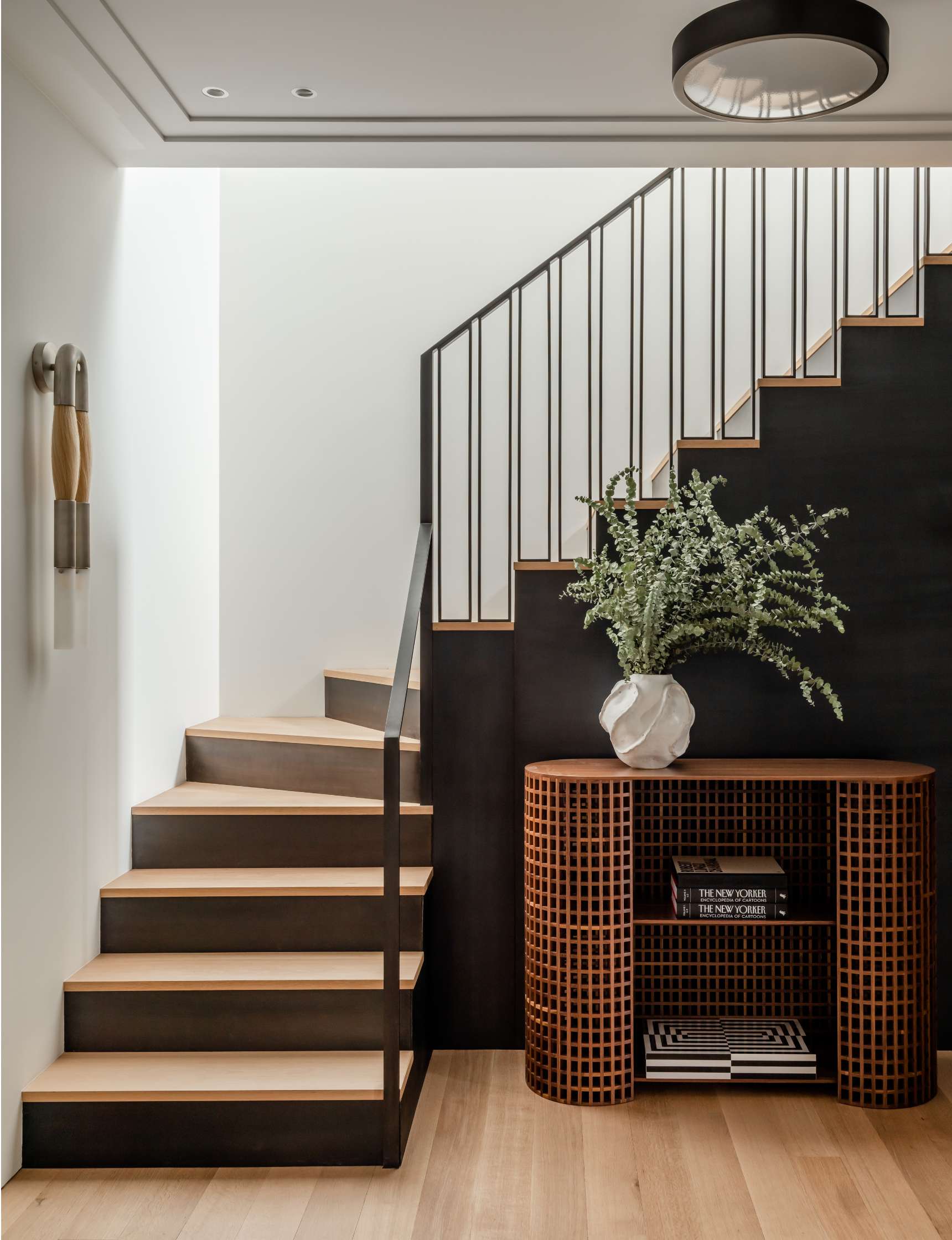 A modern staircase with wooden steps and a metal railing, next to a curvy wooden console holding books and a white vase with green branches. Minimalist wall light fixture above.