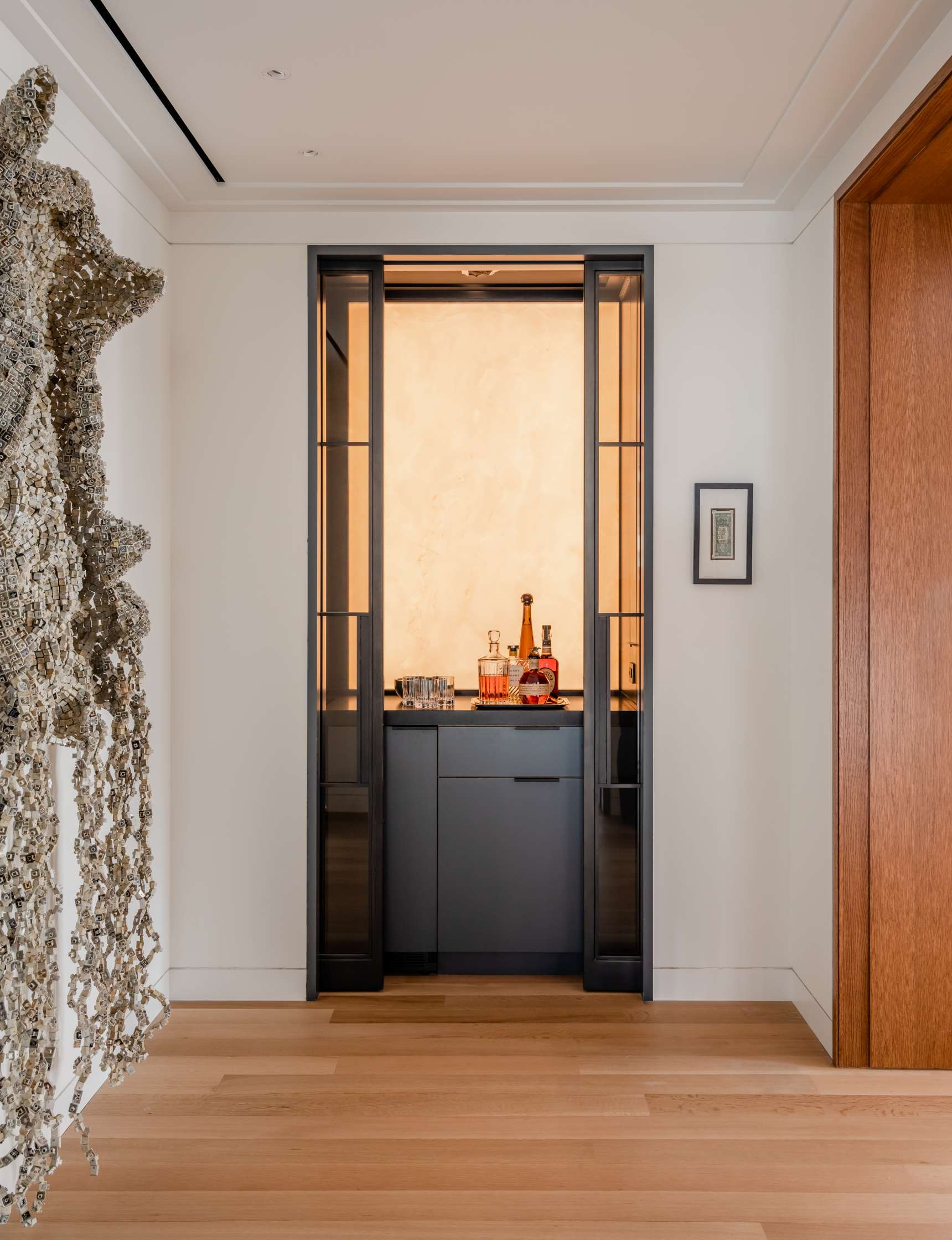 A recessed home bar with glass sliding doors, displaying bottles, glasses, and bar tools on a counter, framed by modern decor including a unique wall sculpture and a small framed picture.