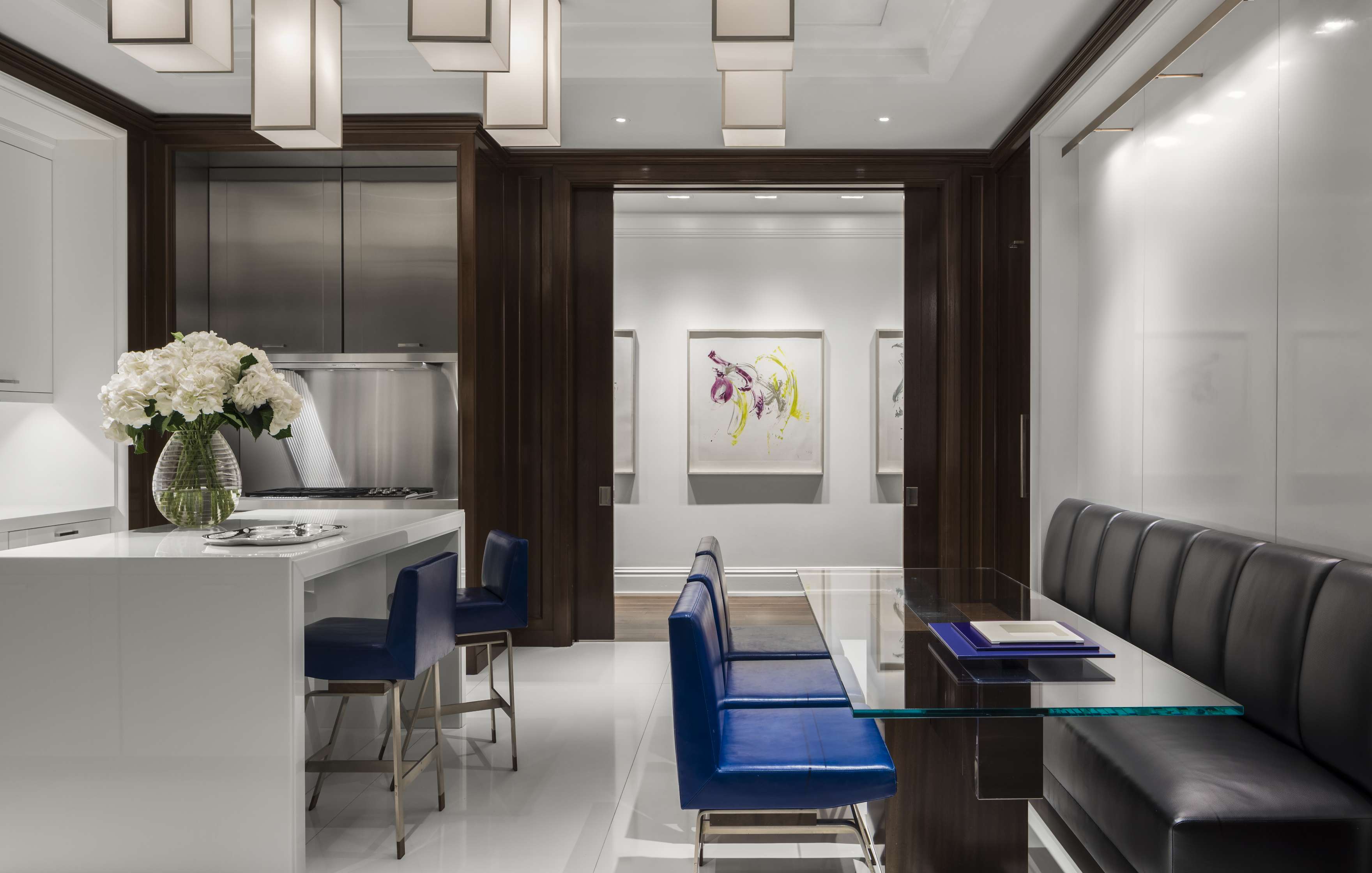 Modern kitchen and dining area with sleek white and dark wood cabinetry, blue padded chairs, glass dining table, white island with flowers, stainless steel appliances, and a framed painting on the wall.