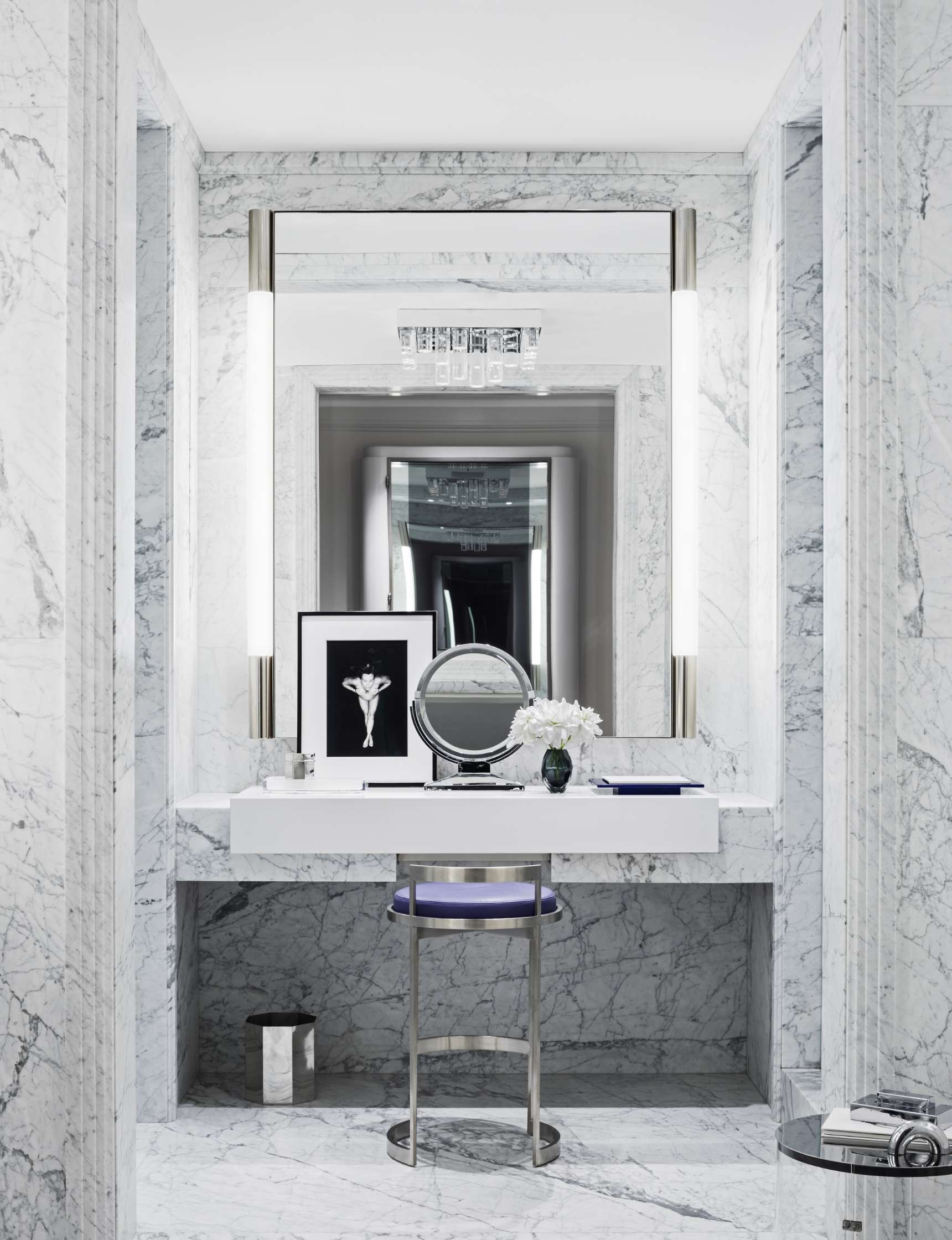 Luxurious marble vanity area with a mirror, bright vertical lights, a small stool, and minimalistic decor including flowers and framed artwork.