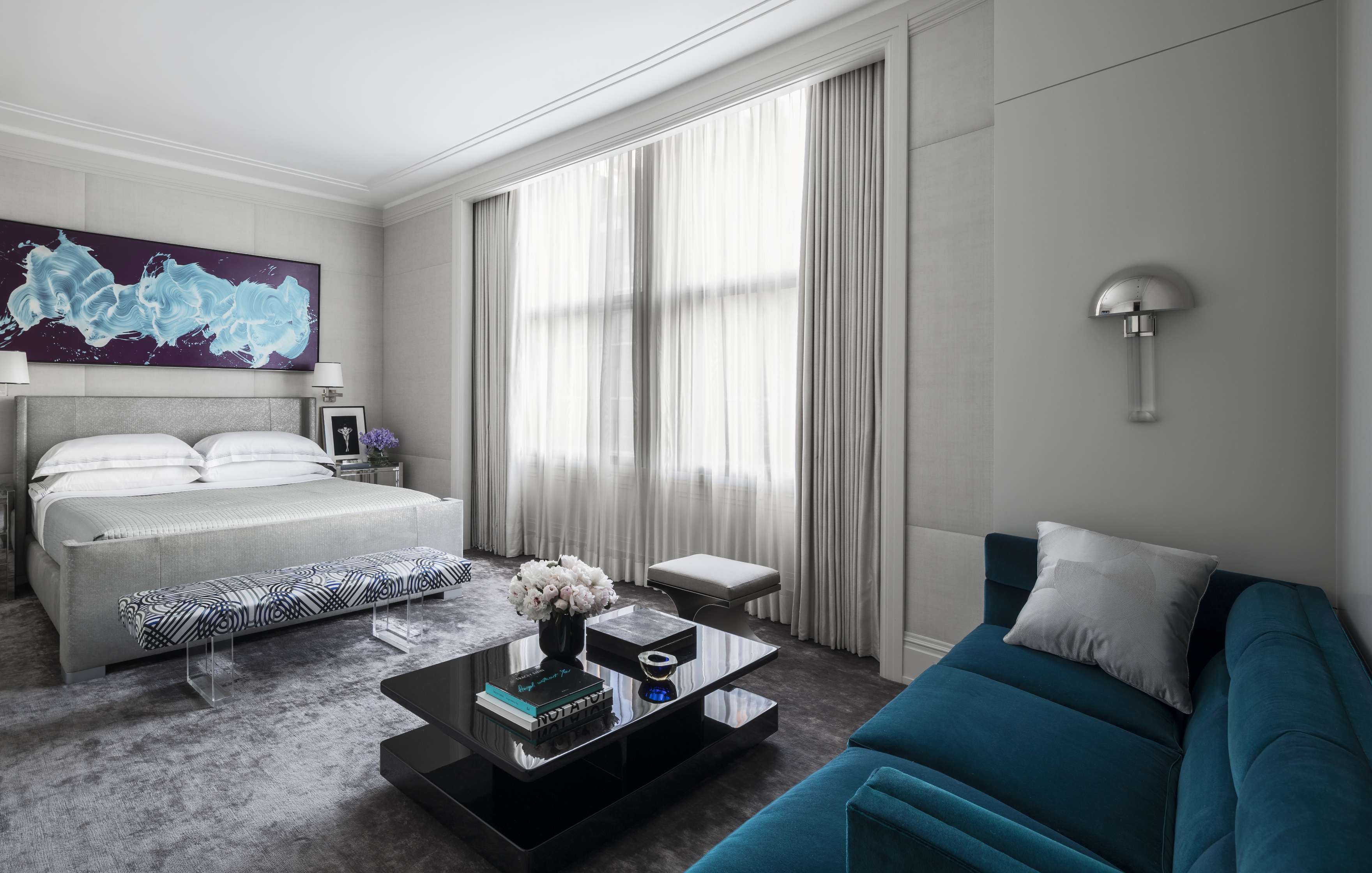 Modern bedroom featuring a bed with white linens, a blue sofa, a sleek black coffee table, an abstract painting, large window with curtains, and a wall-mounted lamp.