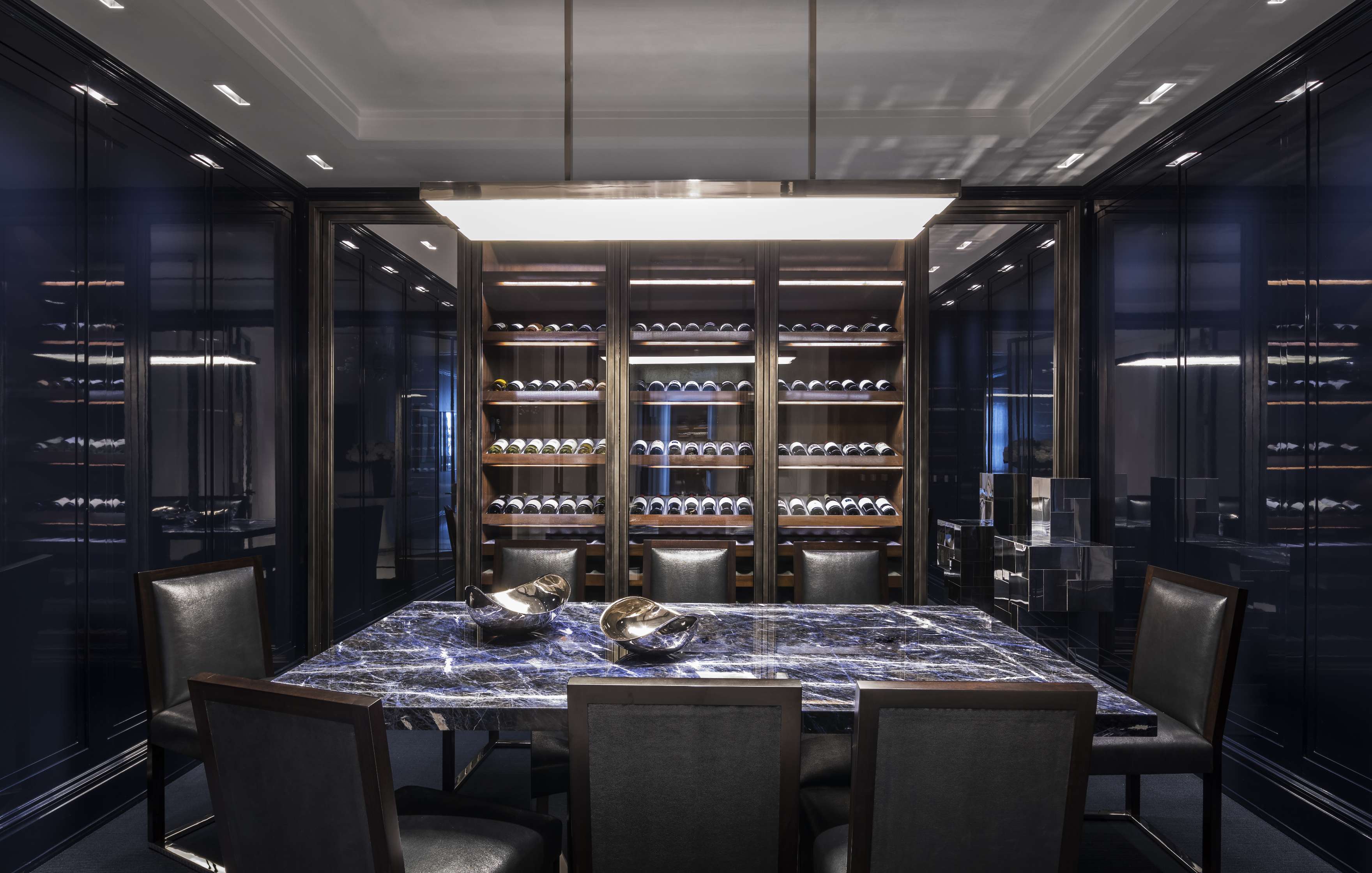 A modern dining room features a large marble table, six black chairs, a bookshelf filled with wine bottles, and contemporary lighting.