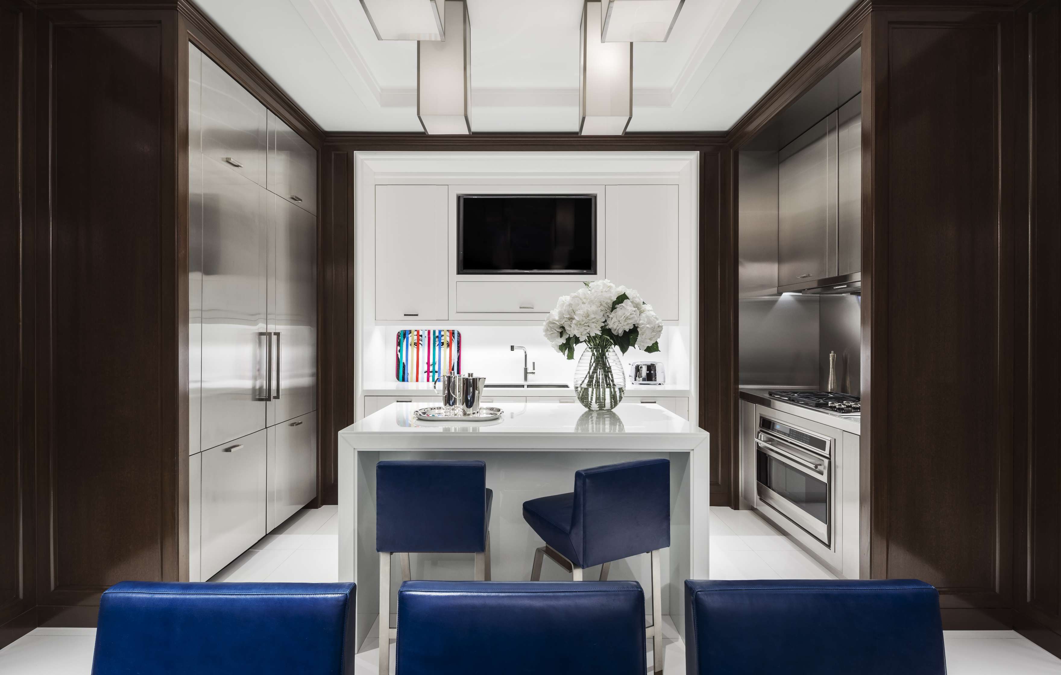 Modern kitchen with dark wood cabinets, stainless steel refrigerator, and oven. A white island with a vase of flowers and blue barstools sits in the center. Ceiling lights illuminate the room.