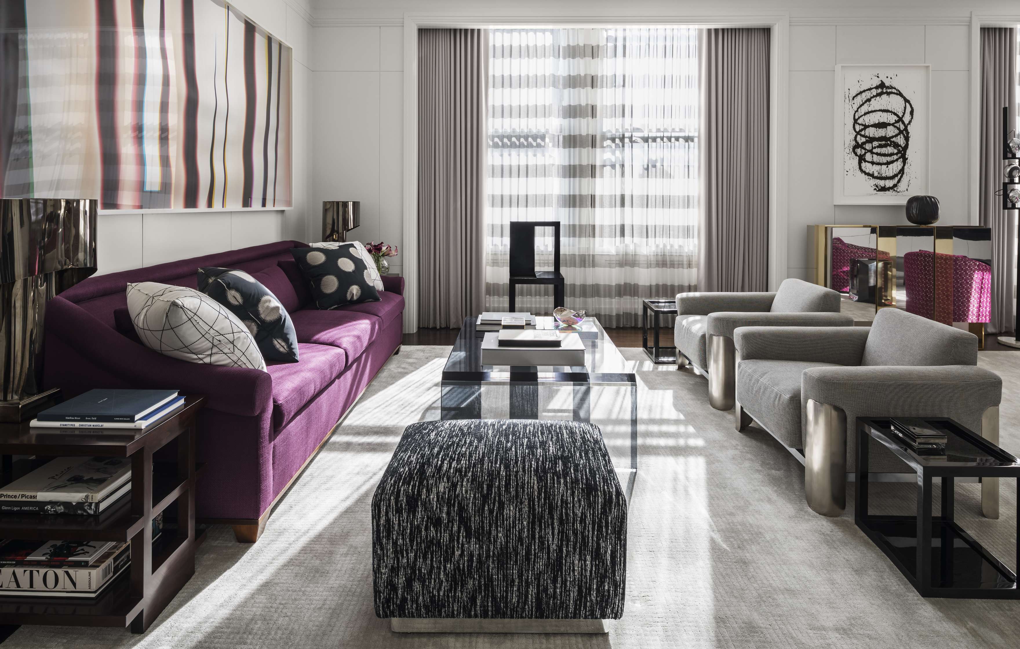 A modern living room with a purple sofa, grey armchairs, a patterned ottoman, a large abstract painting, and floor-to-ceiling windows with sheer drapes.