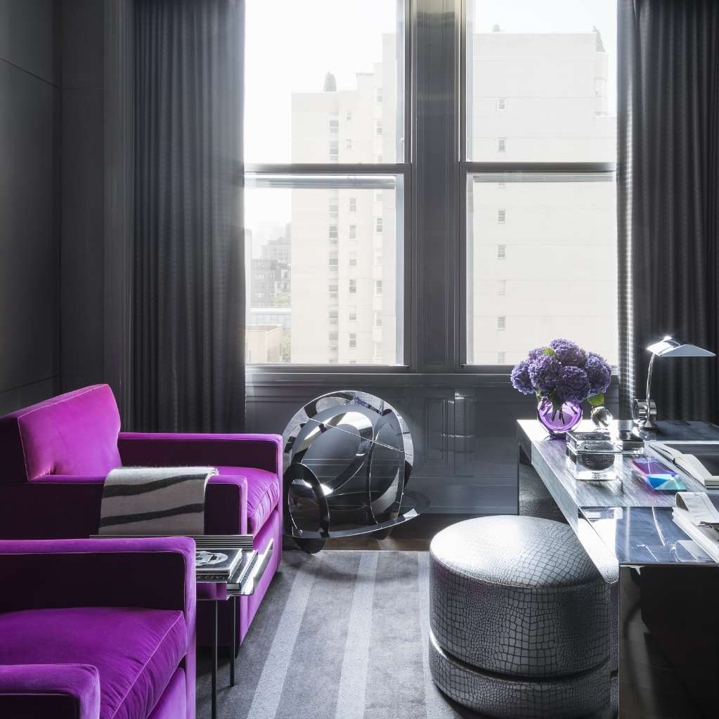 A modern office with a purple chair, a round ottoman, a glass desk with a lamp, books, and a vase of flowers, next to a large window with city buildings visible outside.