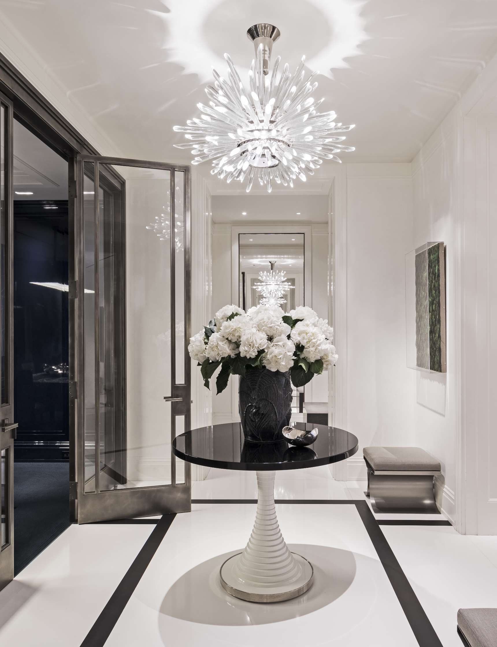 A sleek hallway features a round black table with a vase of white flowers, under a modern chandelier with a starburst design.