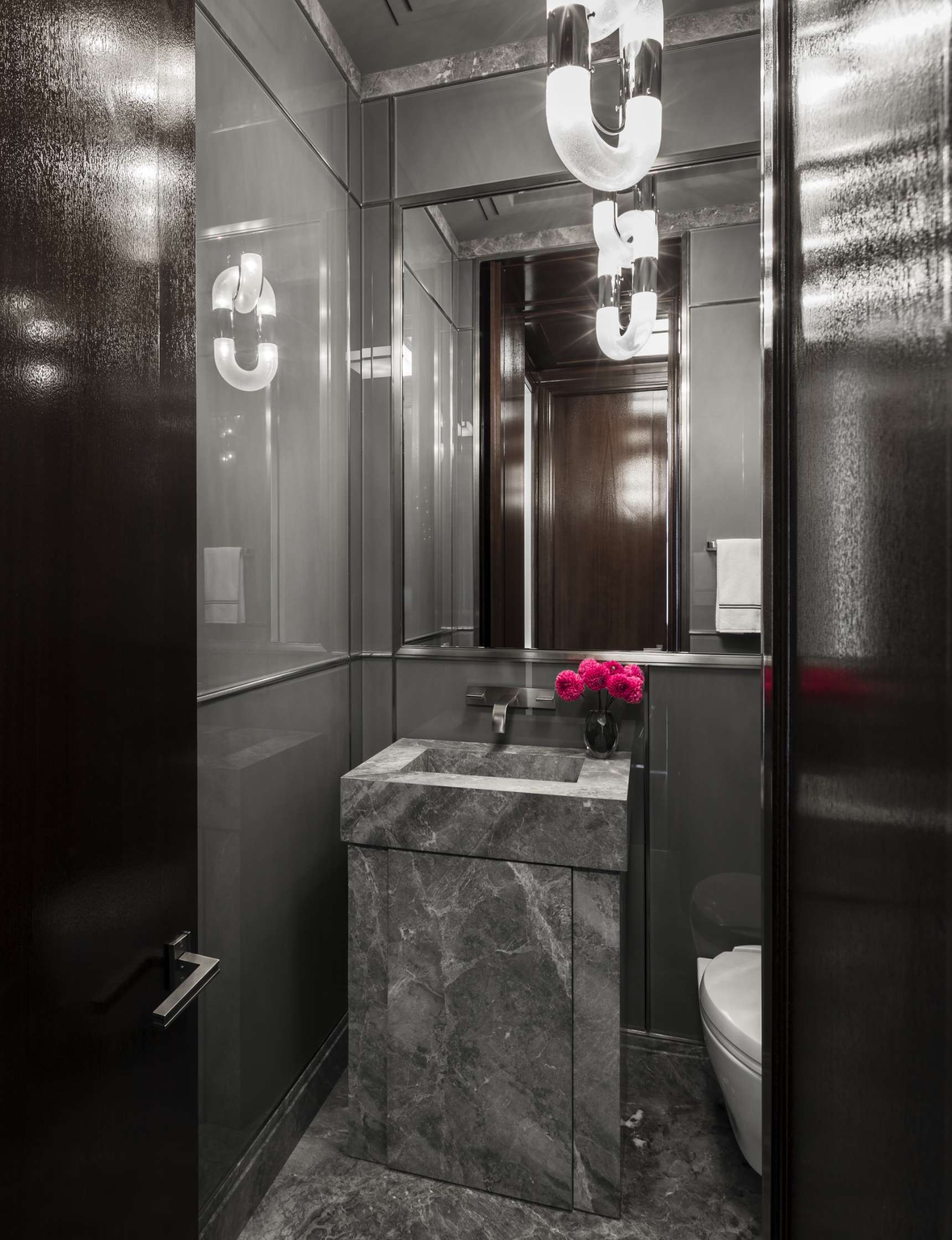 A modern, small bathroom with dark marble walls and floor, a rectangular marble sink, a mirror, and a wall-mounted light fixture. There are pink flowers on the sink and a white towel on the rack.