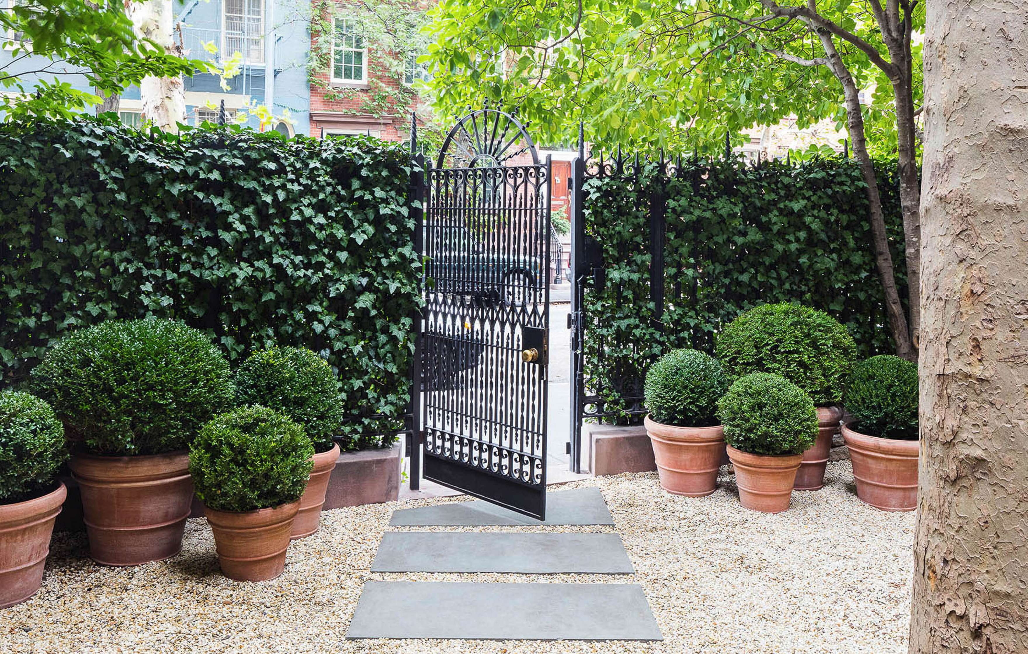 An iron gate is surrounded by green leafy hedges and potted plants, leading to a street. Stone pathways are set in gravel leading to the gate. Trees and buildings are visible beyond the gate.