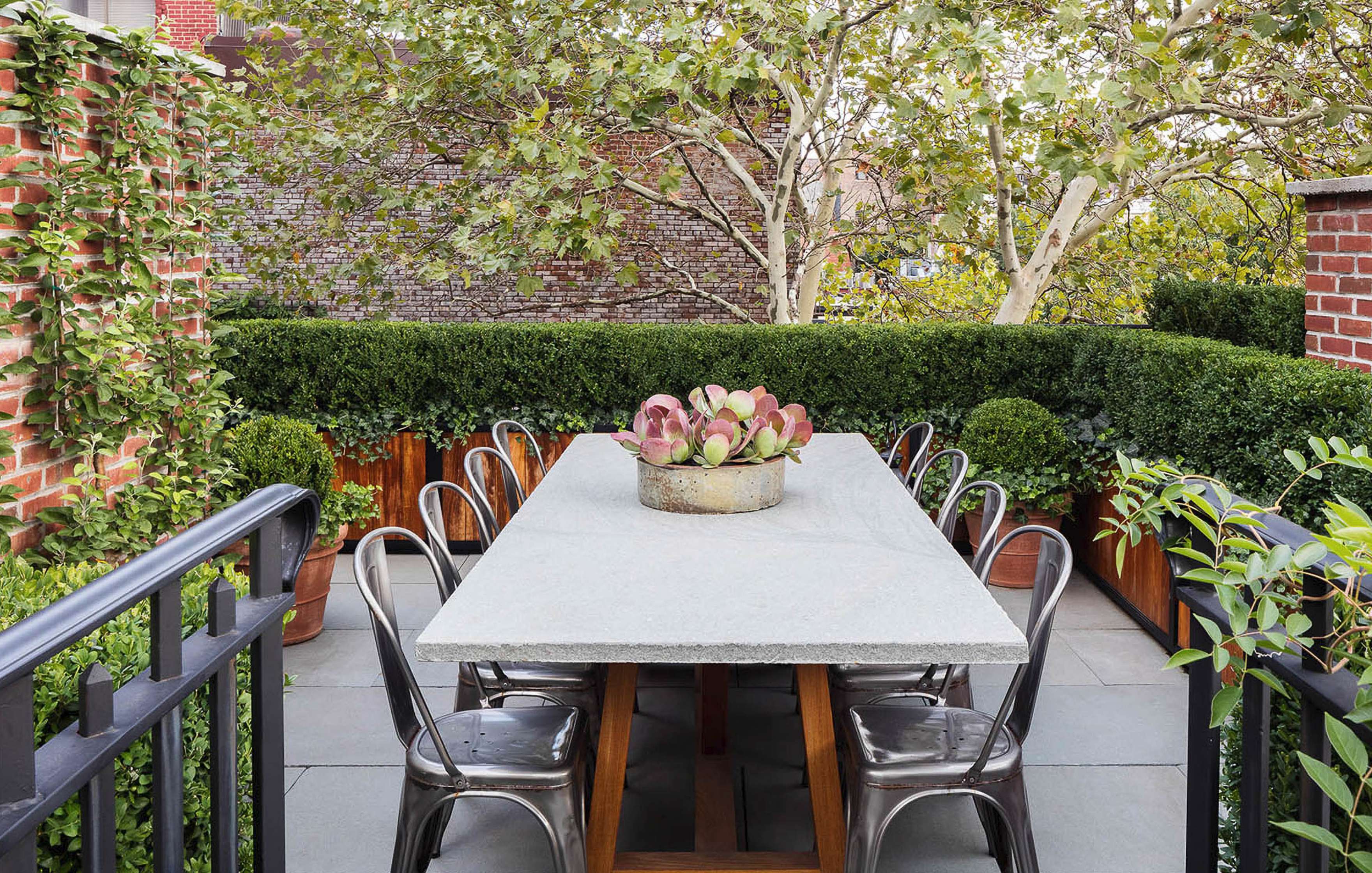 Outdoor dining area with a stone table, surrounded by metal chairs. The table features a center succulent arrangement, and the space is enclosed by lush greenery and brick walls.