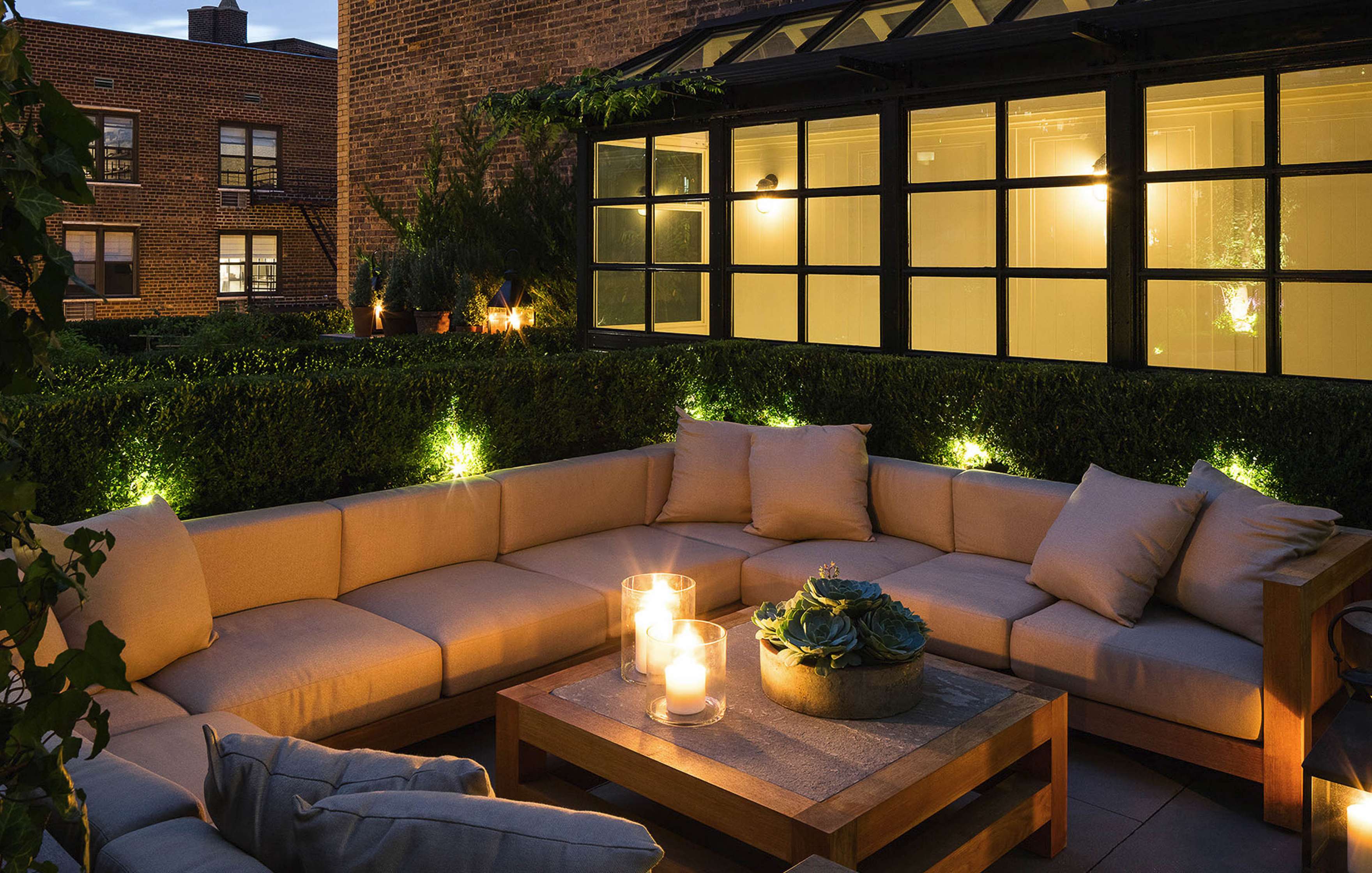 An outdoor patio at dusk with beige cushioned seating, a wooden coffee table holding candles, and plants along the perimeter. Warm lighting illuminates the space.