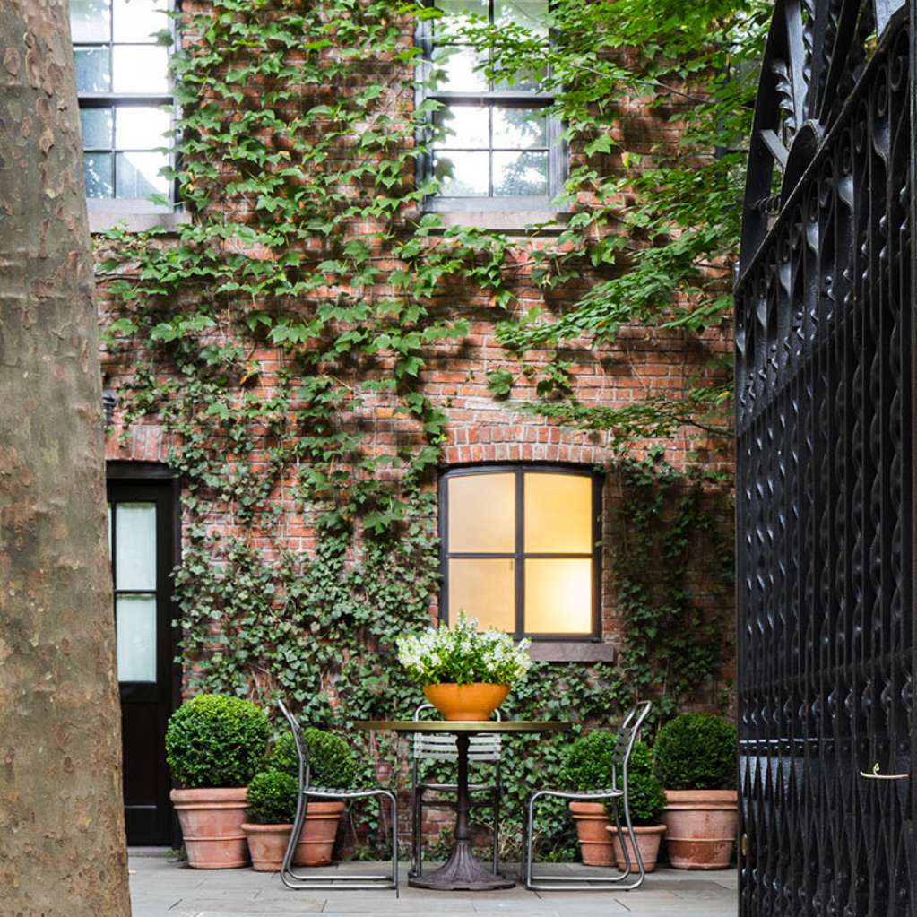 A cozy outdoor patio area with ivy-covered brick wall, a small table with a potted plant, two chairs, and surrounding potted greenery. A black gate is partially visible on the right.