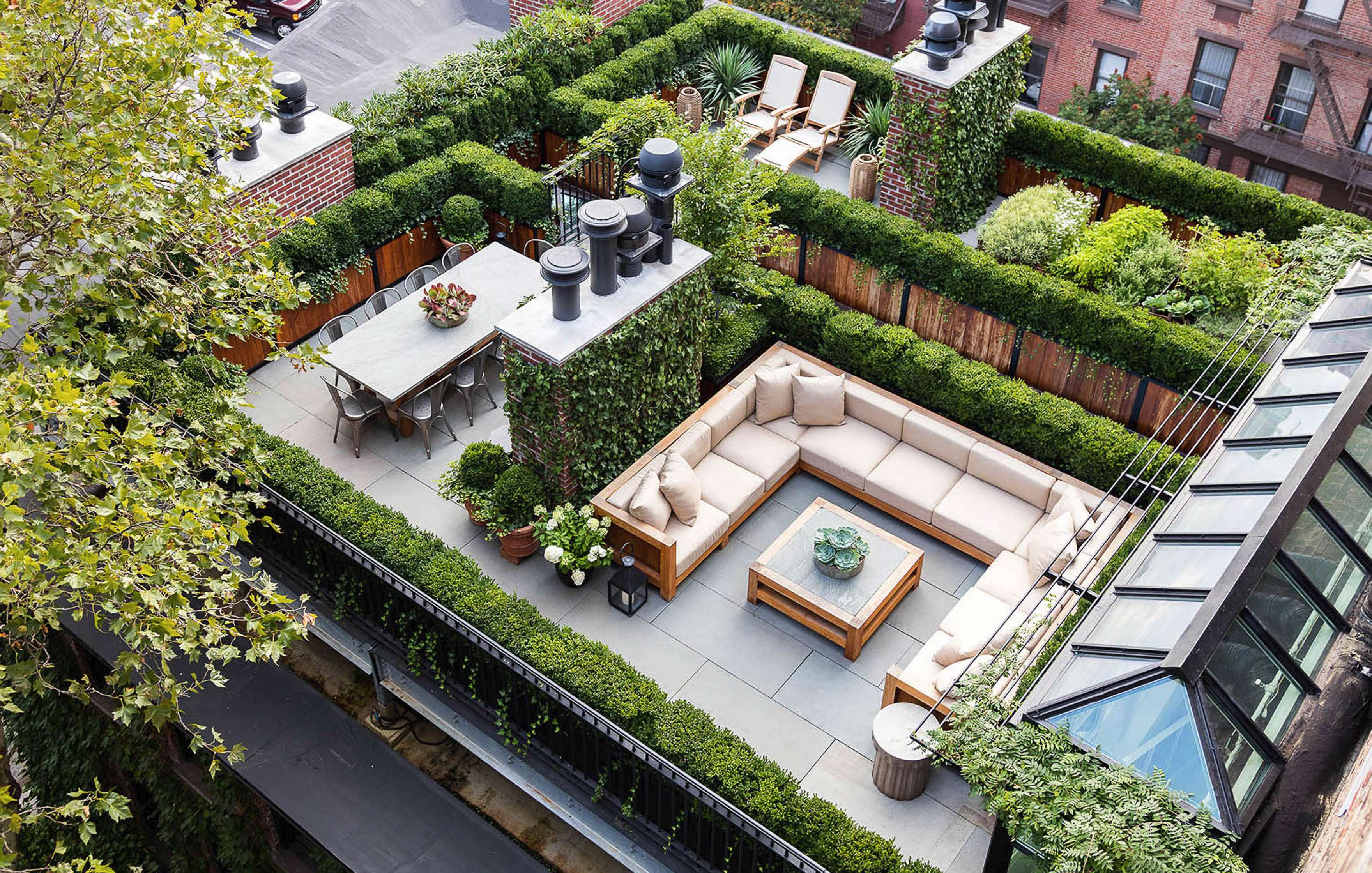 A rooftop garden with sectional seating, a dining table, and greenery walls, surrounded by buildings and trees.