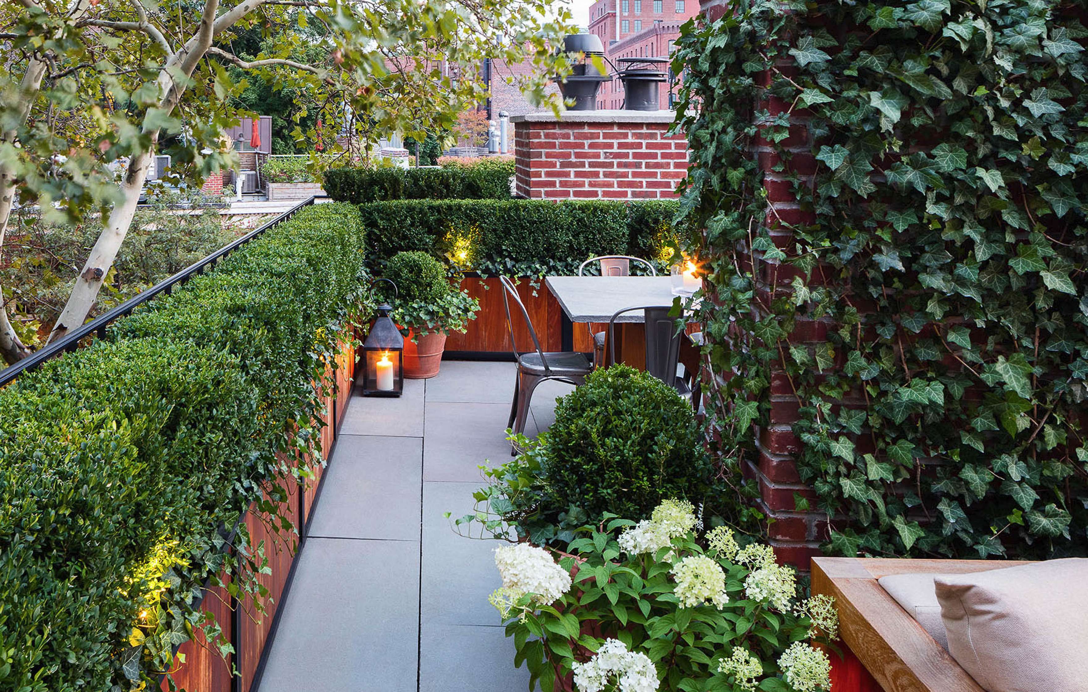 A rooftop patio with a dining table, chairs, and lush green plants. The area is surrounded by hedges, ivy-clad walls, and lanterns for lighting.