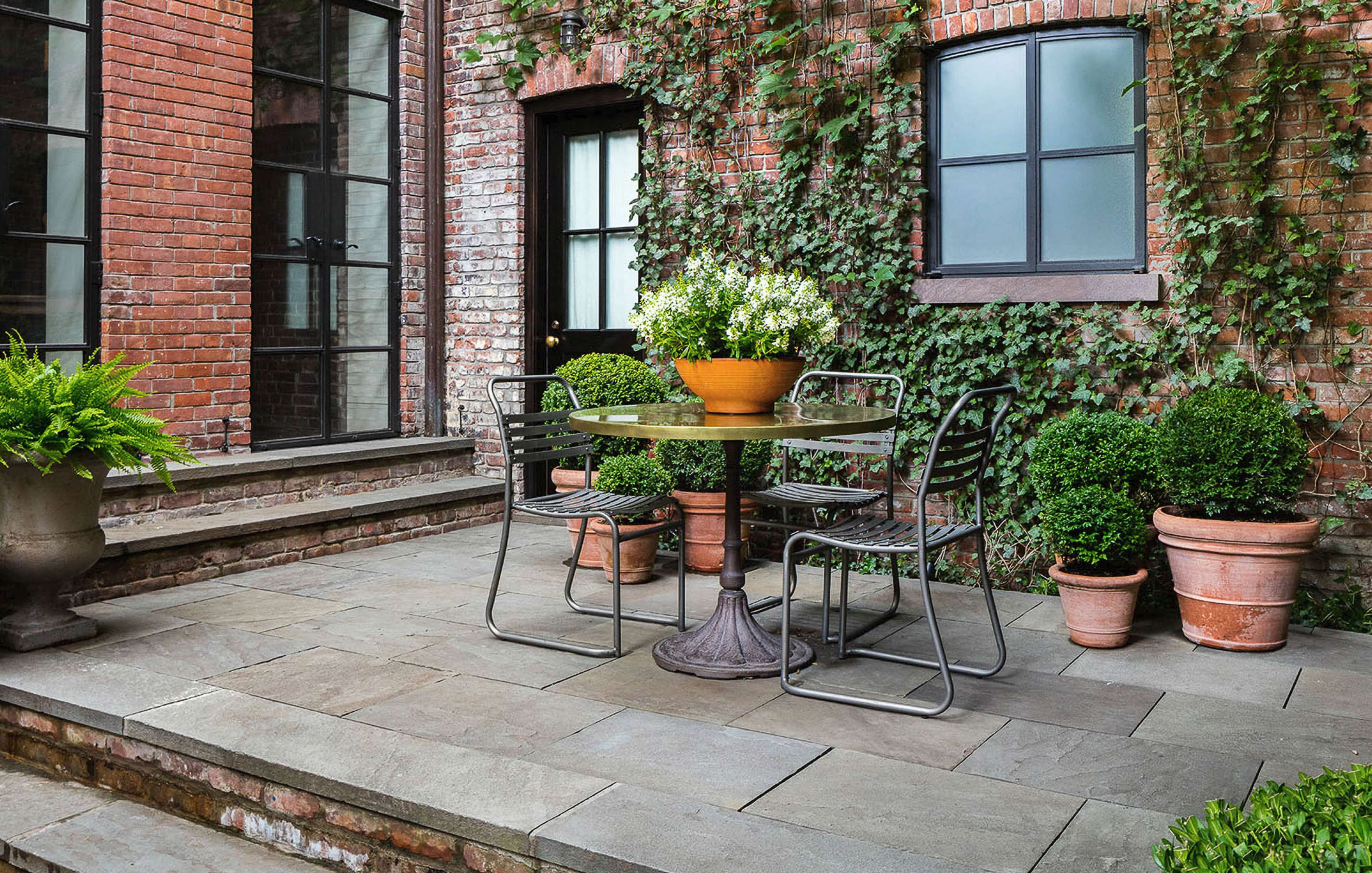 A small outdoor patio with a round table and three chairs, surrounded by potted plants and ivy climbing a brick wall.