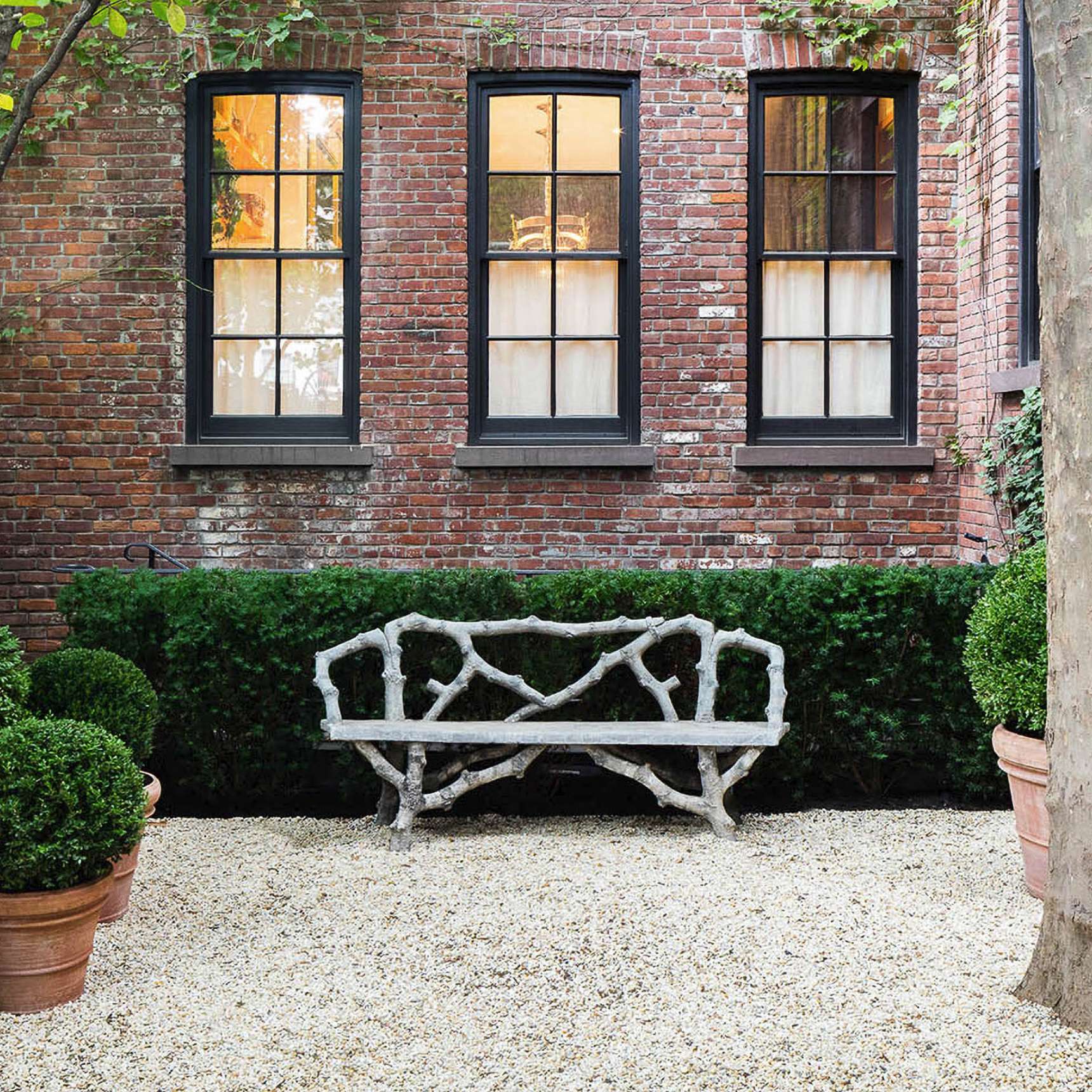 A stone bench is placed in front of a neatly trimmed green hedge against a brick building with three rectangular windows. Potted plants are arranged on a gravel surface beside the bench.