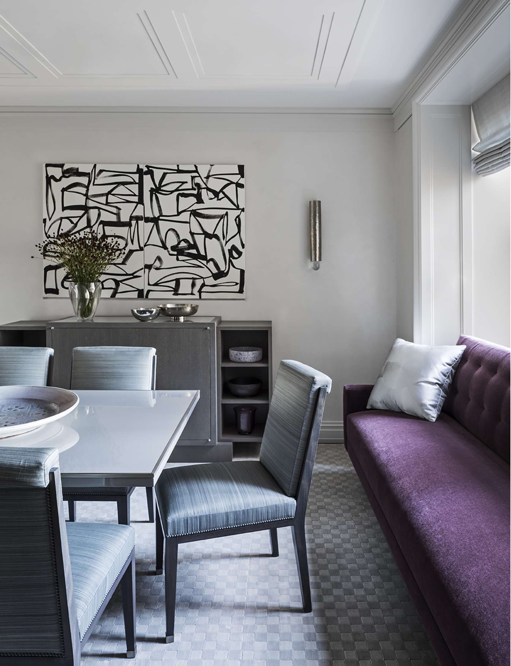 Modern dining room with a purple sofa, gray dining chairs, a white table, and abstract black-and-white wall art. A sideboard with decorative items and a vase of flowers sits against the back wall.