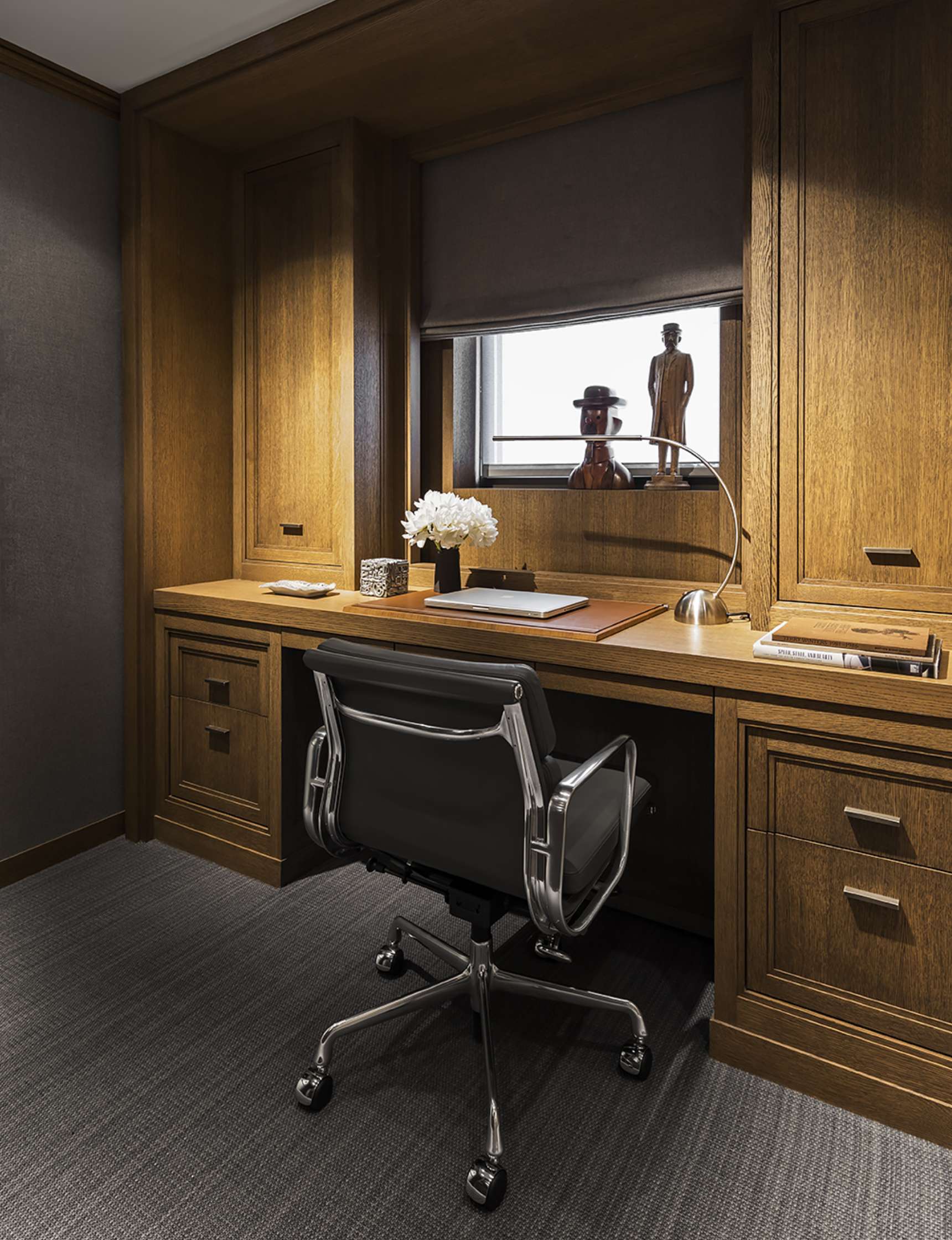 A small, neatly arranged home office features a wooden desk with a laptop, lamp, flowers, and decorative items. An ergonomic chair is positioned in front of the desk.