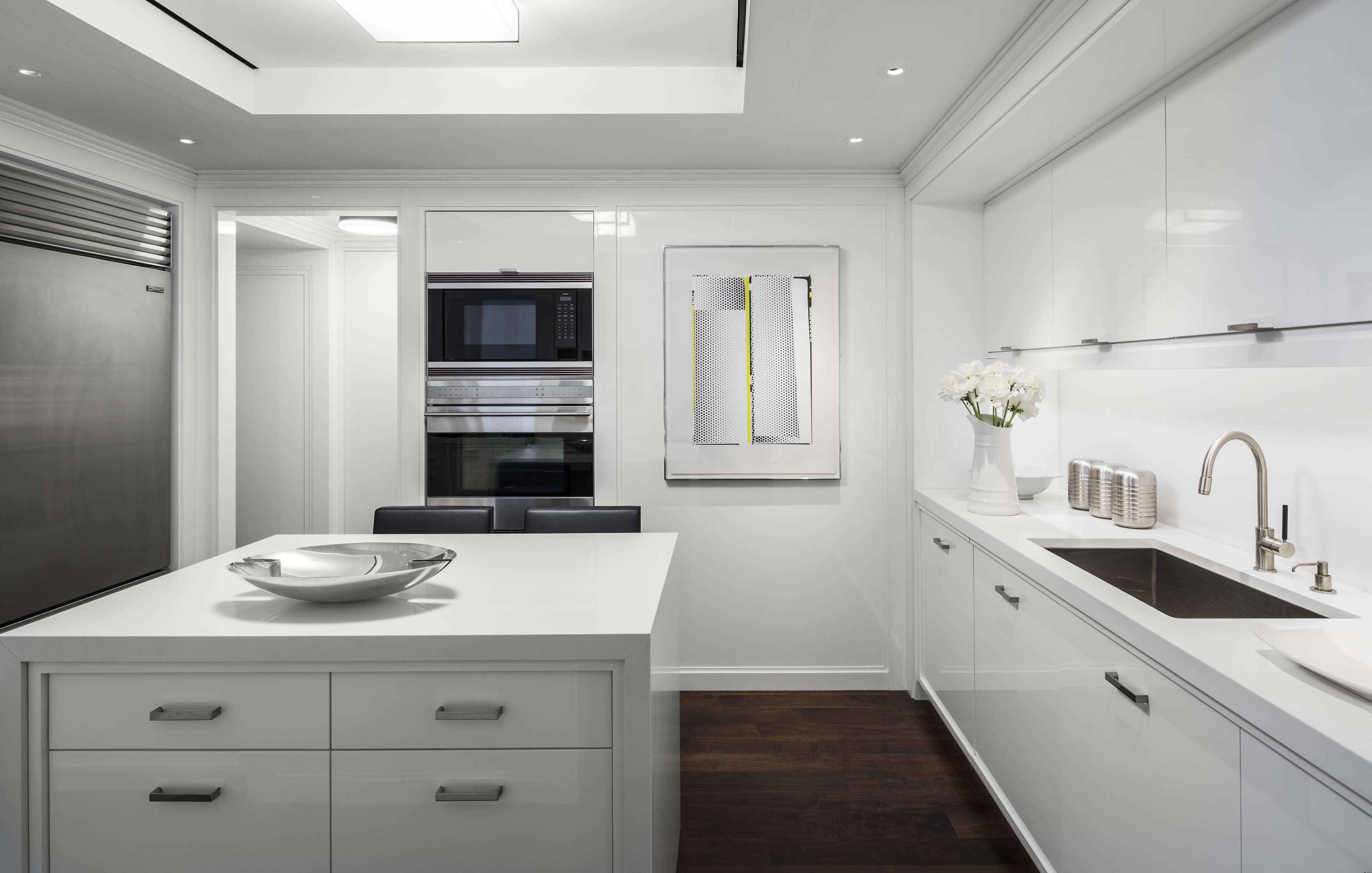 A modern kitchen with white cabinets, stainless steel appliances, a countertop with a bowl, a sink with a faucet, a wall-mounted artwork, and a bouquet of white flowers in a vase.