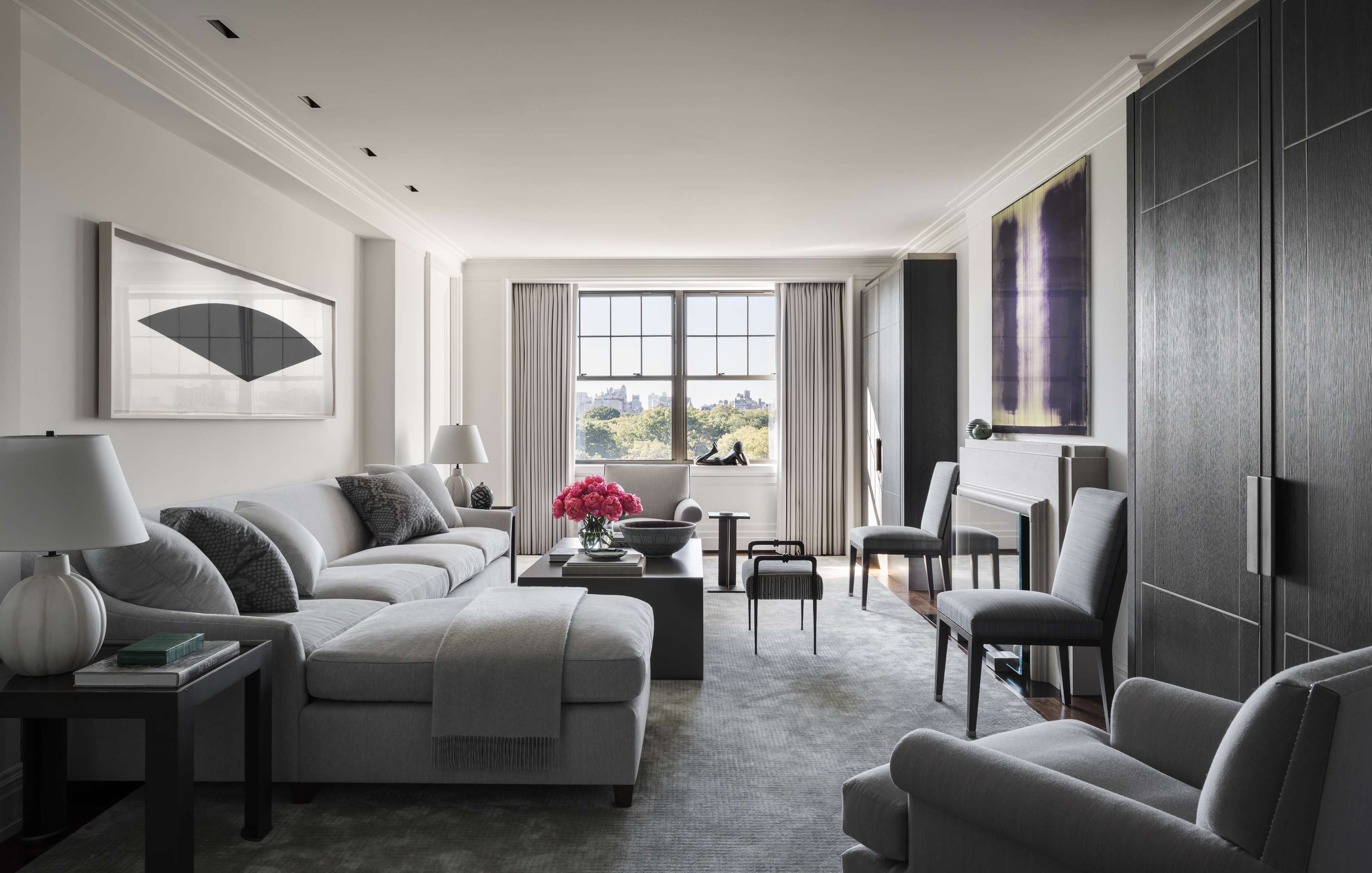 A modern living room with neutral colors, featuring a sectional sofa, armchairs, a coffee table with pink flowers, framed artwork, floor-to-ceiling windows, and built-in cabinets.