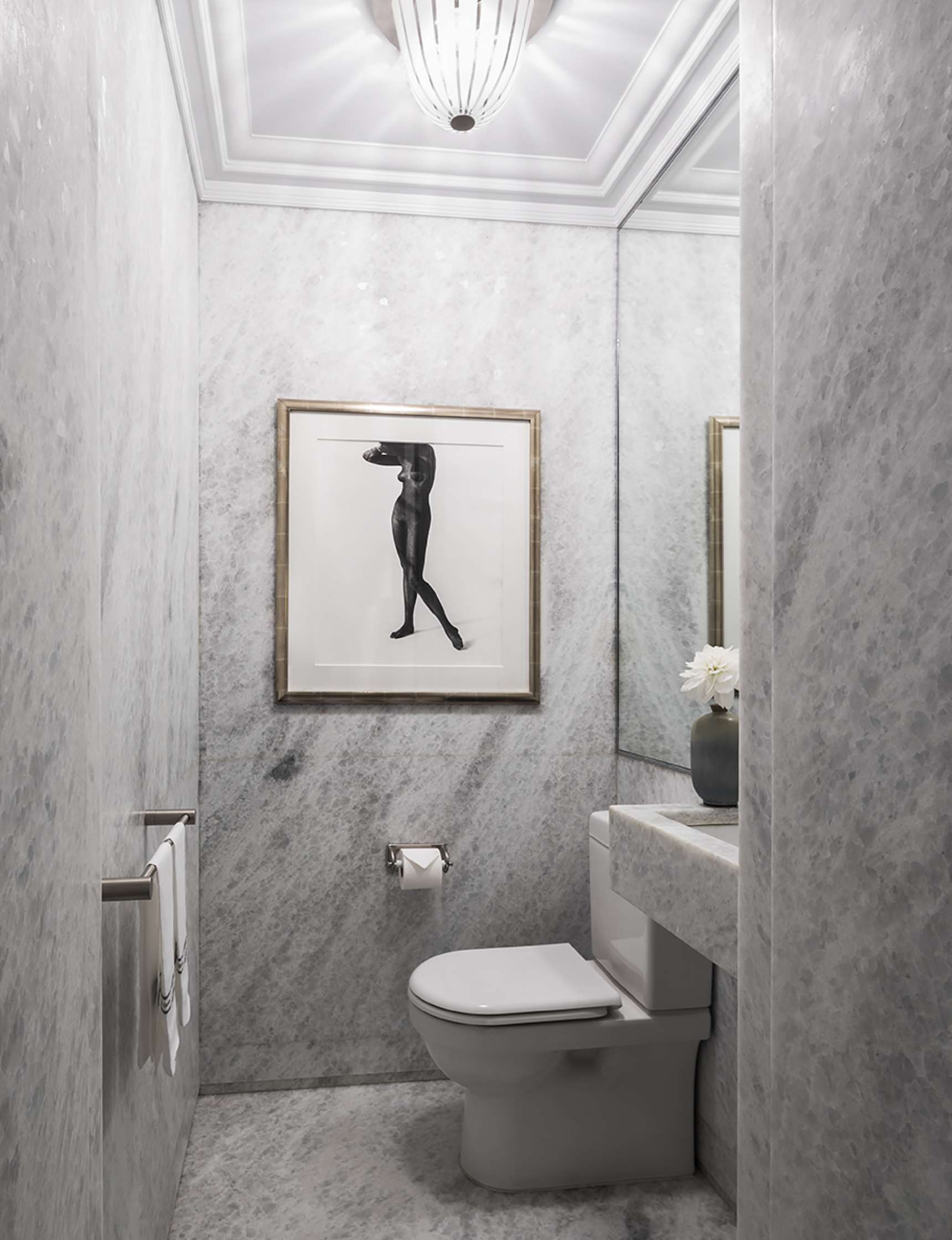 A small marble bathroom with a framed black-and-white artwork, a toilet, a towel rack with white towels, a small vanity with a vase of flowers, and an overhead light fixture.