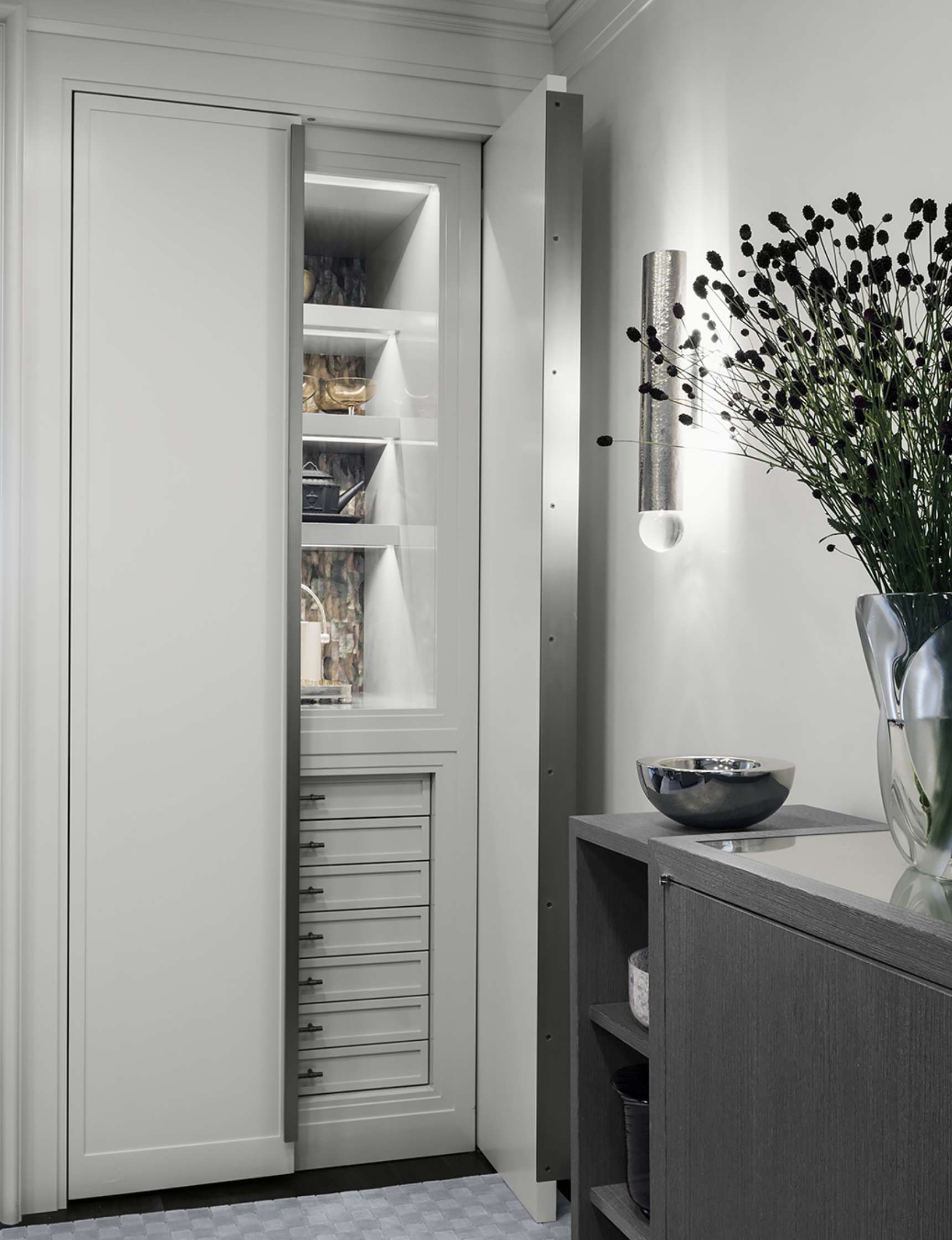 A minimalist, built-in white cabinet with sliding doors, internal shelves containing decorative items, and a gray side table with a glass vase and a small bowl, illuminated by a wall-mounted light fixture.