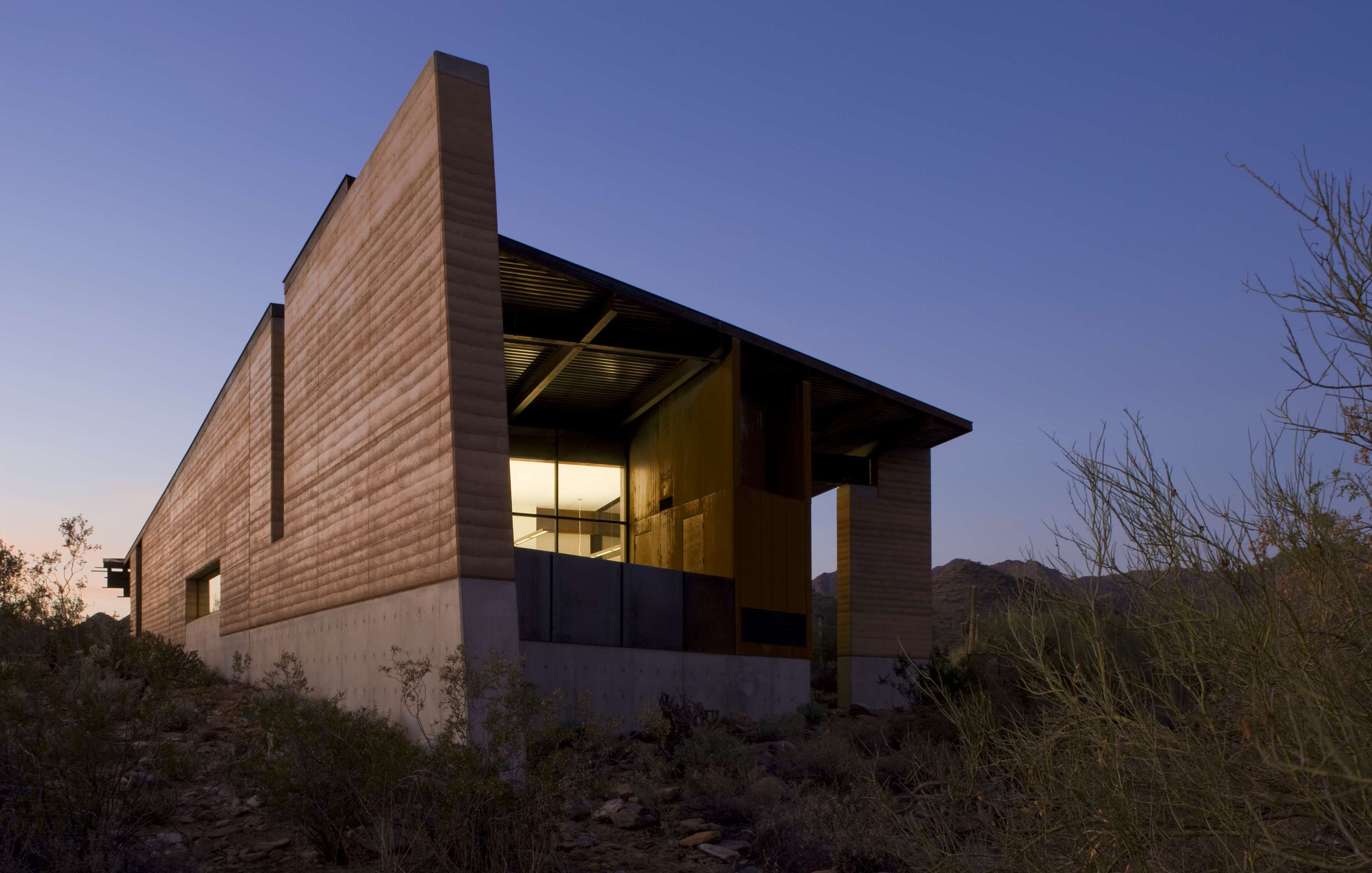 A modern, minimalist building with large windows, set in a desert landscape at dusk, surrounded by sparse vegetation.