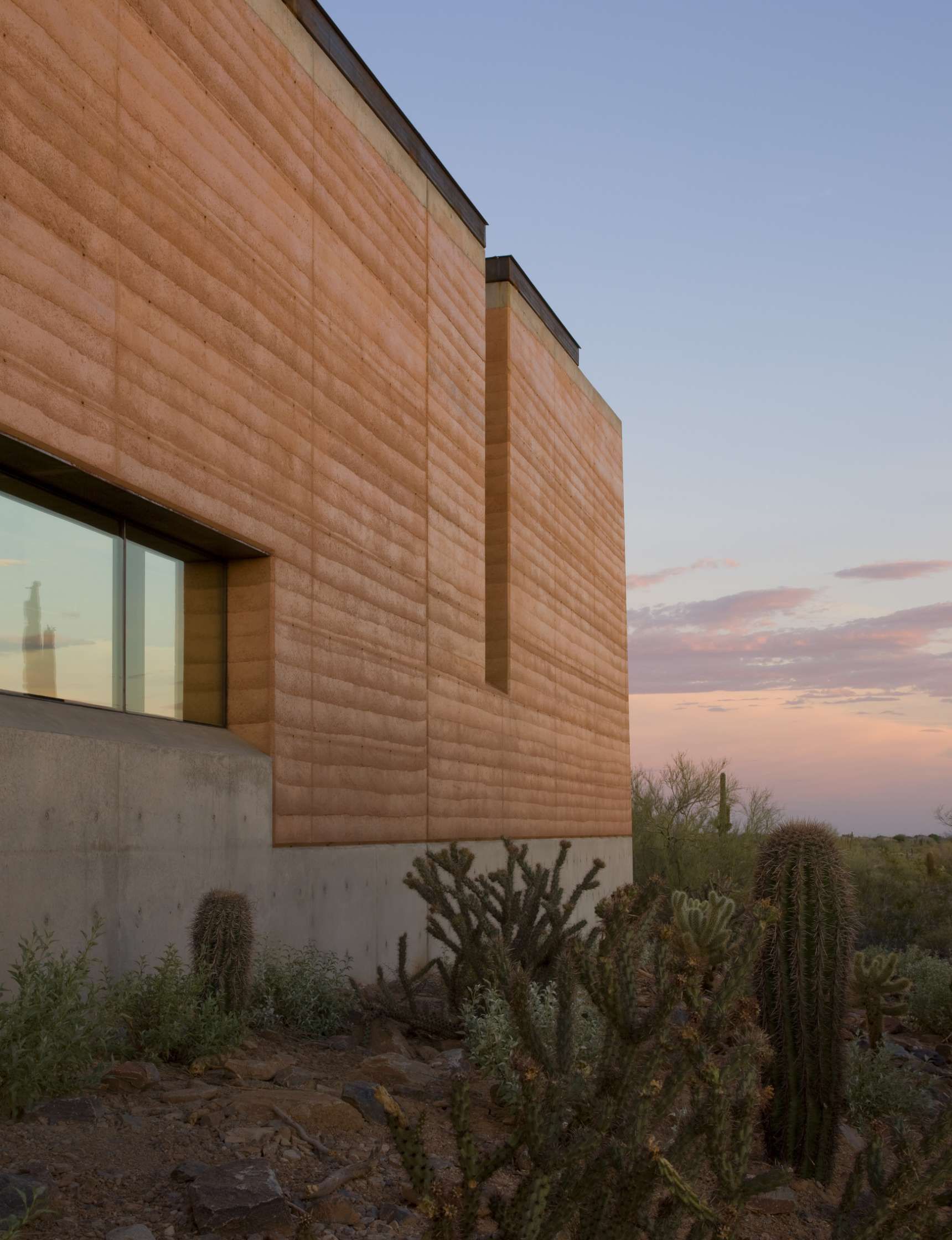 Modern building with earthy-toned walls and a large window, surrounded by desert plants and cacti, under a twilight sky.