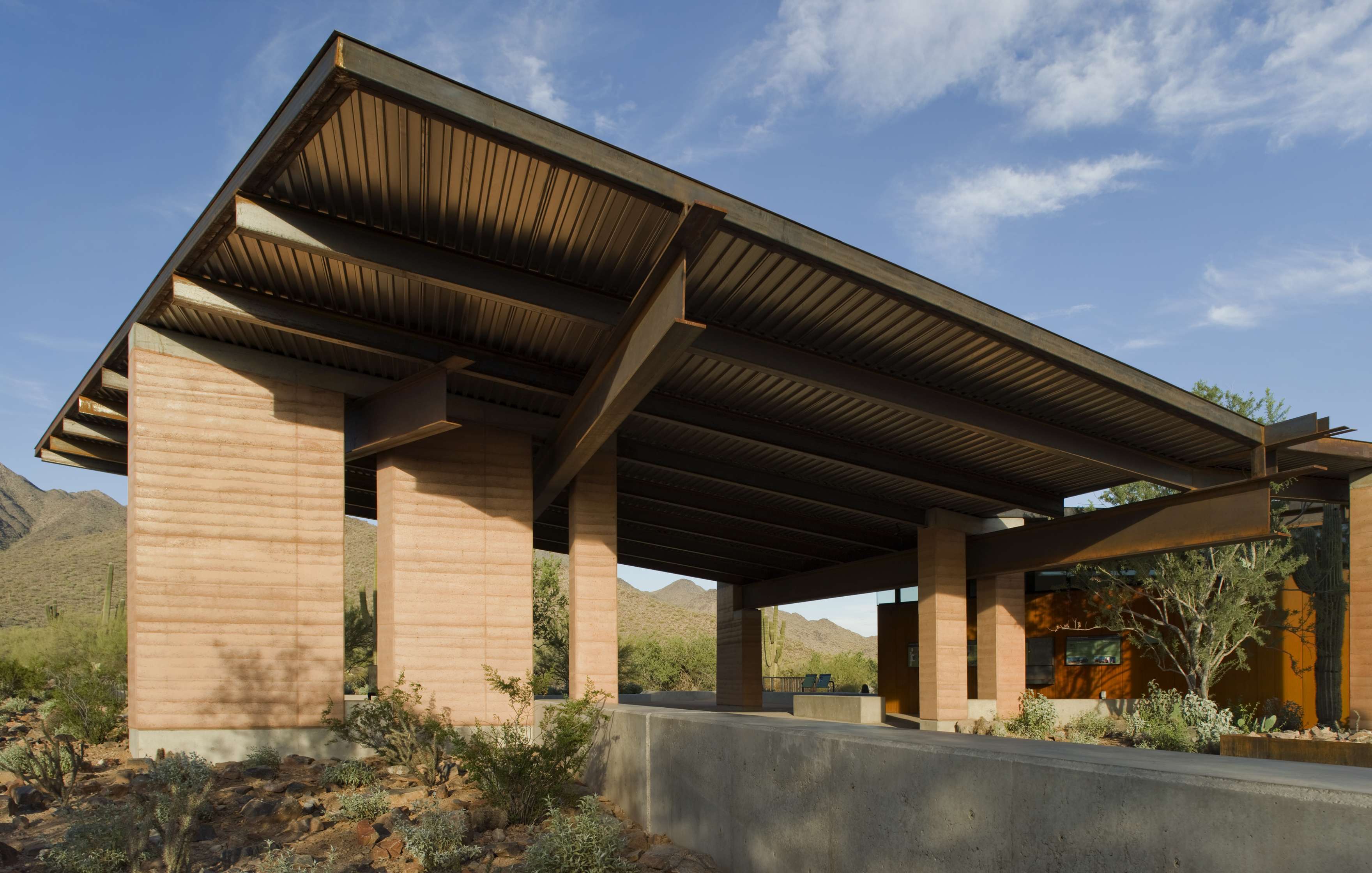 A modern building with a large, flat roof supported by thick pillars and surrounded by a landscaped desert environment under a partly cloudy sky.