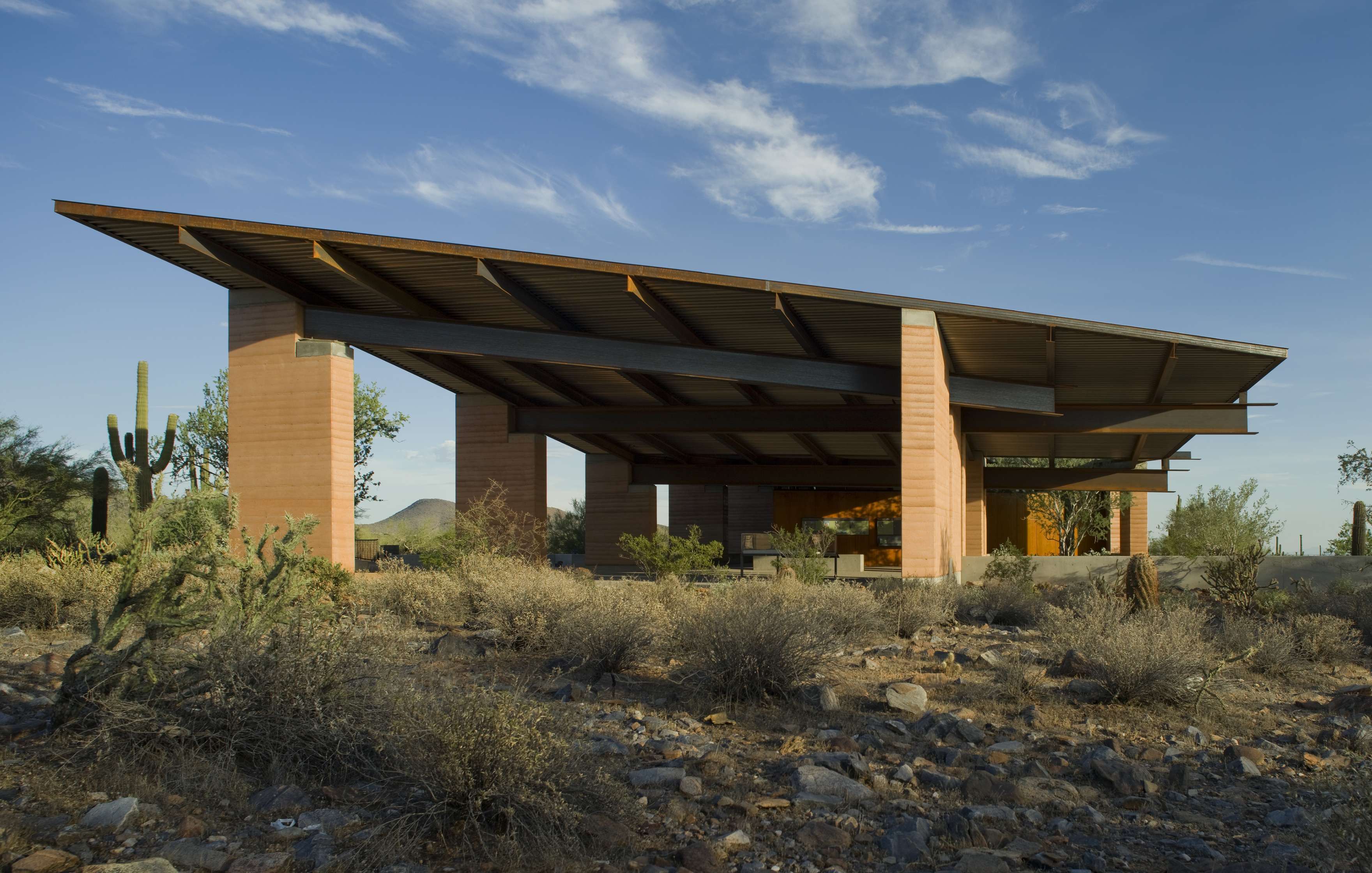 A modern house with a large overhanging roof set in a desert landscape with cacti and dry shrubs under a blue sky with scattered clouds.