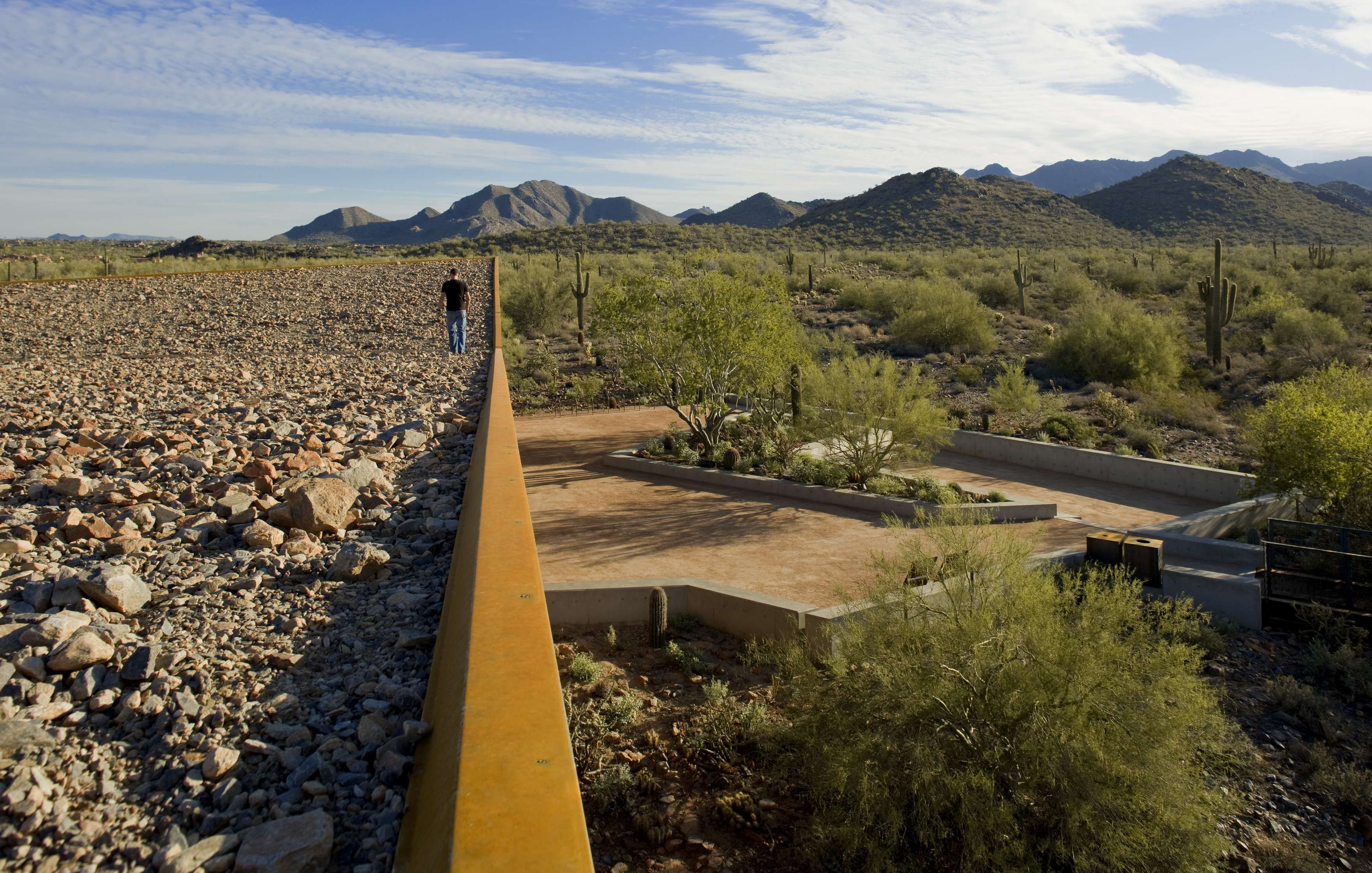 A person walks along a rocky path beside a desert landscape with mountains in the background. Cacti and shrubs are scattered around the area.