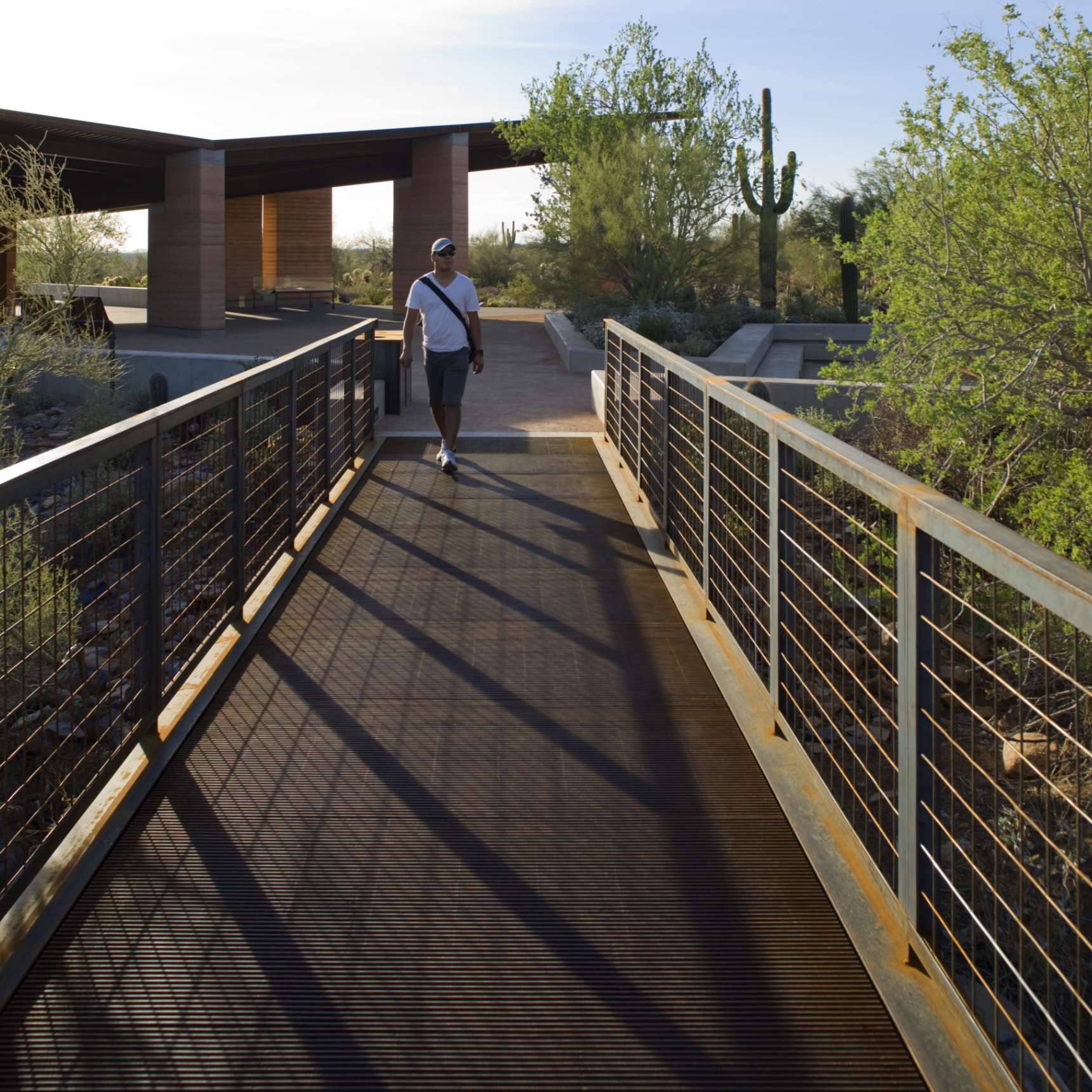 A person walks on a metal bridge with trees, cactus, and a building in the background on a sunny day.